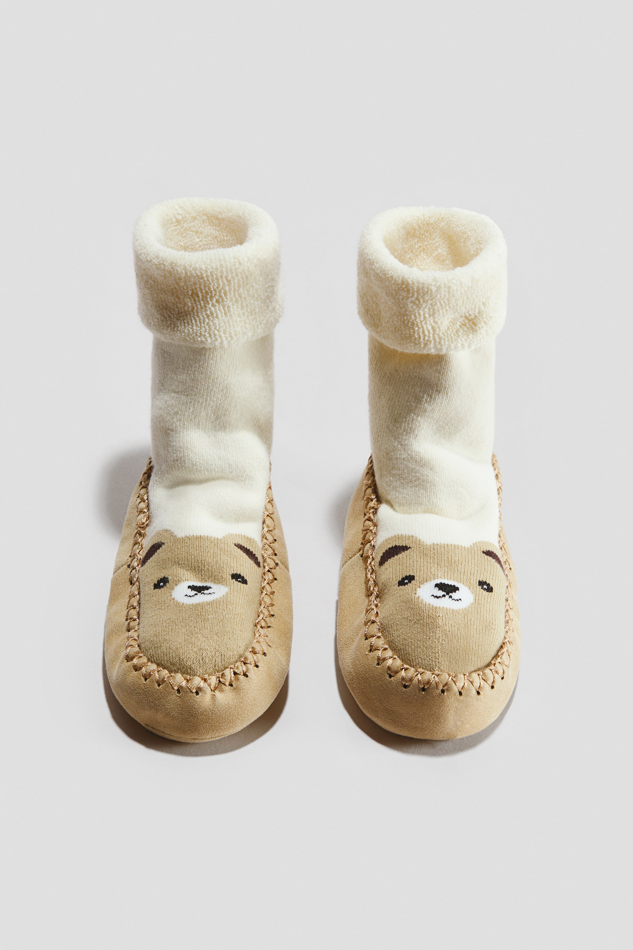 Voir une image plus grande: Une paire de chaussettes-chaussons pour bébé présente une partie supérieure couleur crème avec un revers moelleux replié. Le dessous de chausson beige affiche un motif de visage d’ours avec des détails brodés et des coutures de style mocassin sur l’avant.