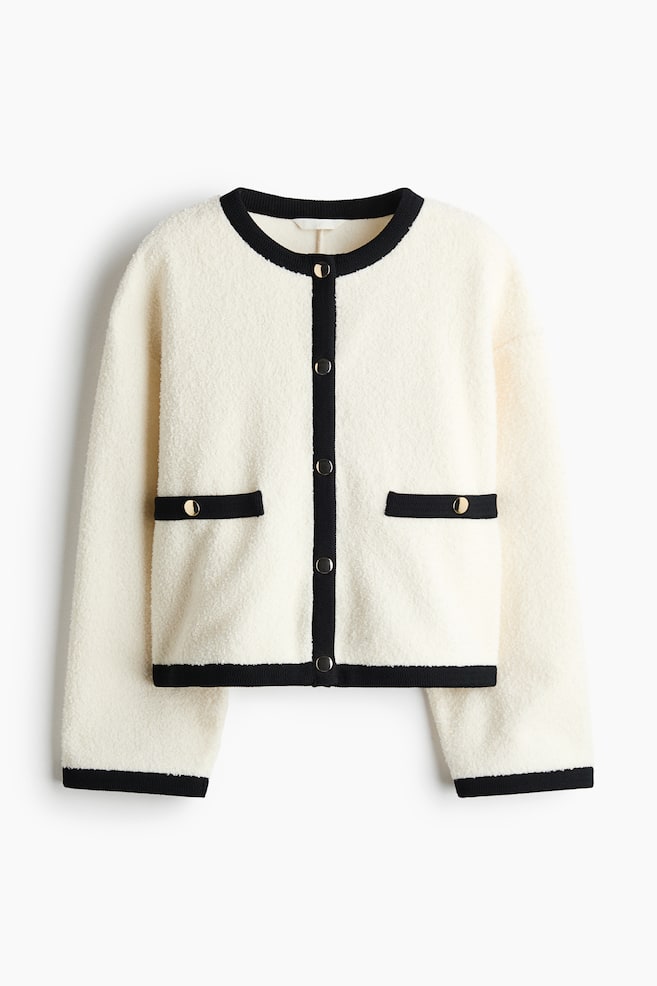A cream-colored teddy jacket is facing front, featuring a relaxed fit, dropped shoulders, and a round neckline. It is displaying contrasting black ribbed trim along the collar, center placket, cuffs, hem, and mock welt pockets, accented by gold-colored metal buttons. - 1