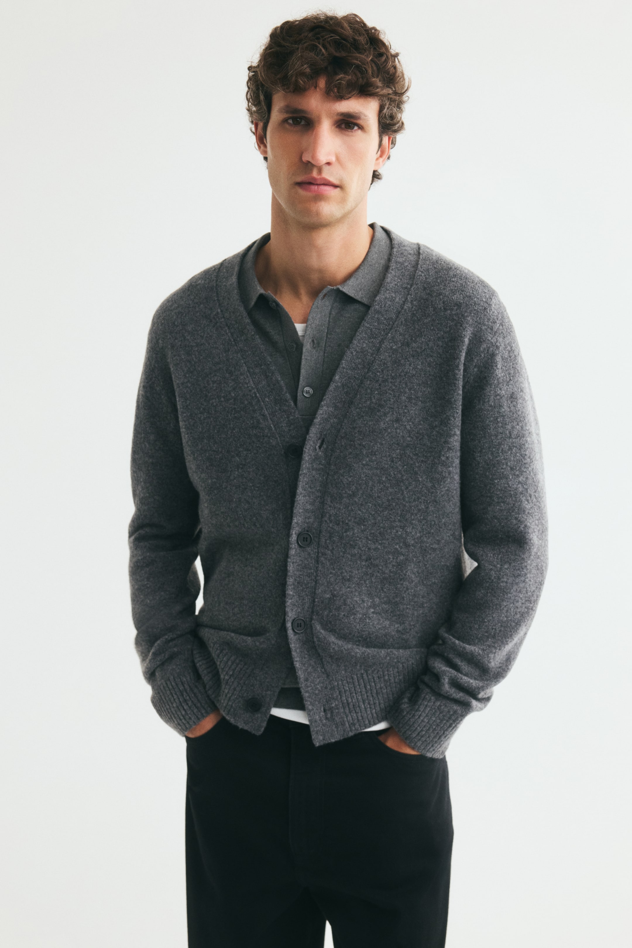 Größeres Bild anzeigen: Ein grauer, weich gestrickter Cardigan mit V-Ausschnitt, Knopfleiste vorne und langen Ärmeln wird vom Model getragen. Der Cardigan ist über einem grauen Poloshirt getragen und mit einer schwarzen Hose kombiniert, während die Hände des Models in den Hosentaschen ruhen.
