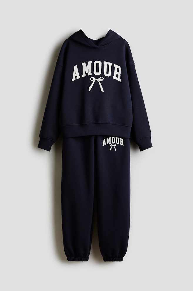 Ein marineblauer zweiteiliger Trainingsanzug präsentiert einen passenden Hoodie und Jogginghosen, beide nach vorne gerichtet. Der Hoodie zeigt 'AMOUR' in weißem Collegeschriftzug mit einer Schleifengrafik darunter, zusammen mit gerippten Bündchen und Saum, während die Jogginghosen denselben Text und die Grafik auf dem oberen linken Bein zeigen und mit elastischen Knöcheln abschließen. - 1