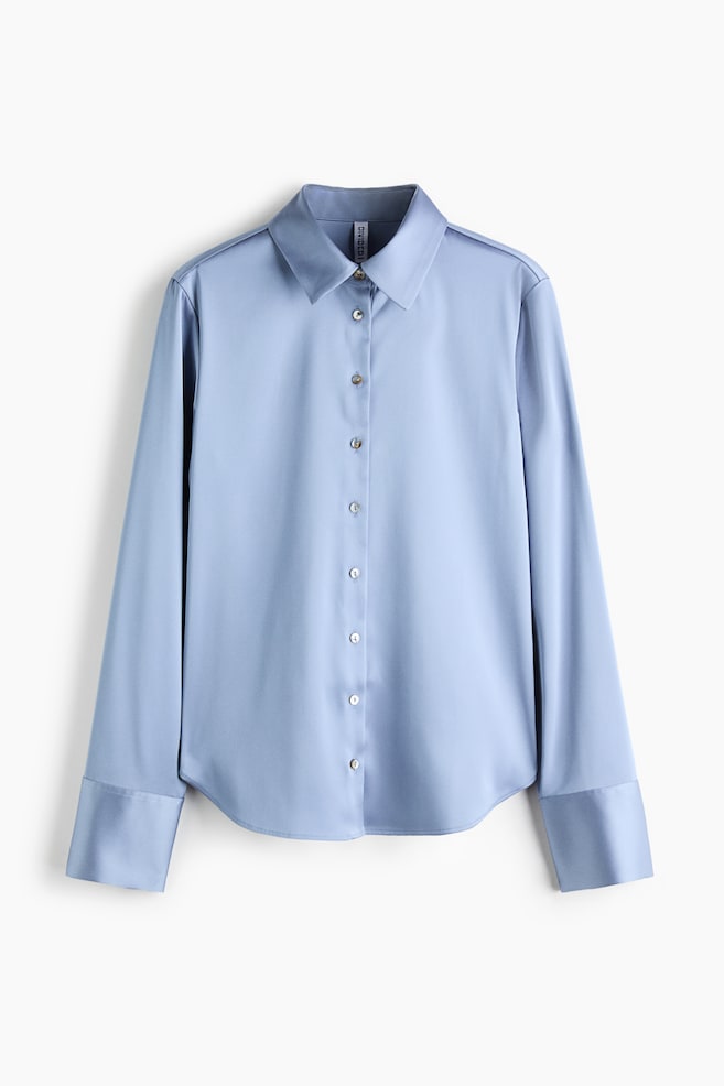 Ein hellblaues, tailliertes Twill-Hemd ist von vorne zu sehen und verfügt über einen spitzen Kragen sowie eine mittig verlaufende Knopfleiste mit kleinen, perlmuttartigen Knöpfen. Die langen Ãrmel enden in breiten, strukturierten Manschetten, und das Hemd ist mit einem sanft abgerundeten Saum gestaltet. - 1