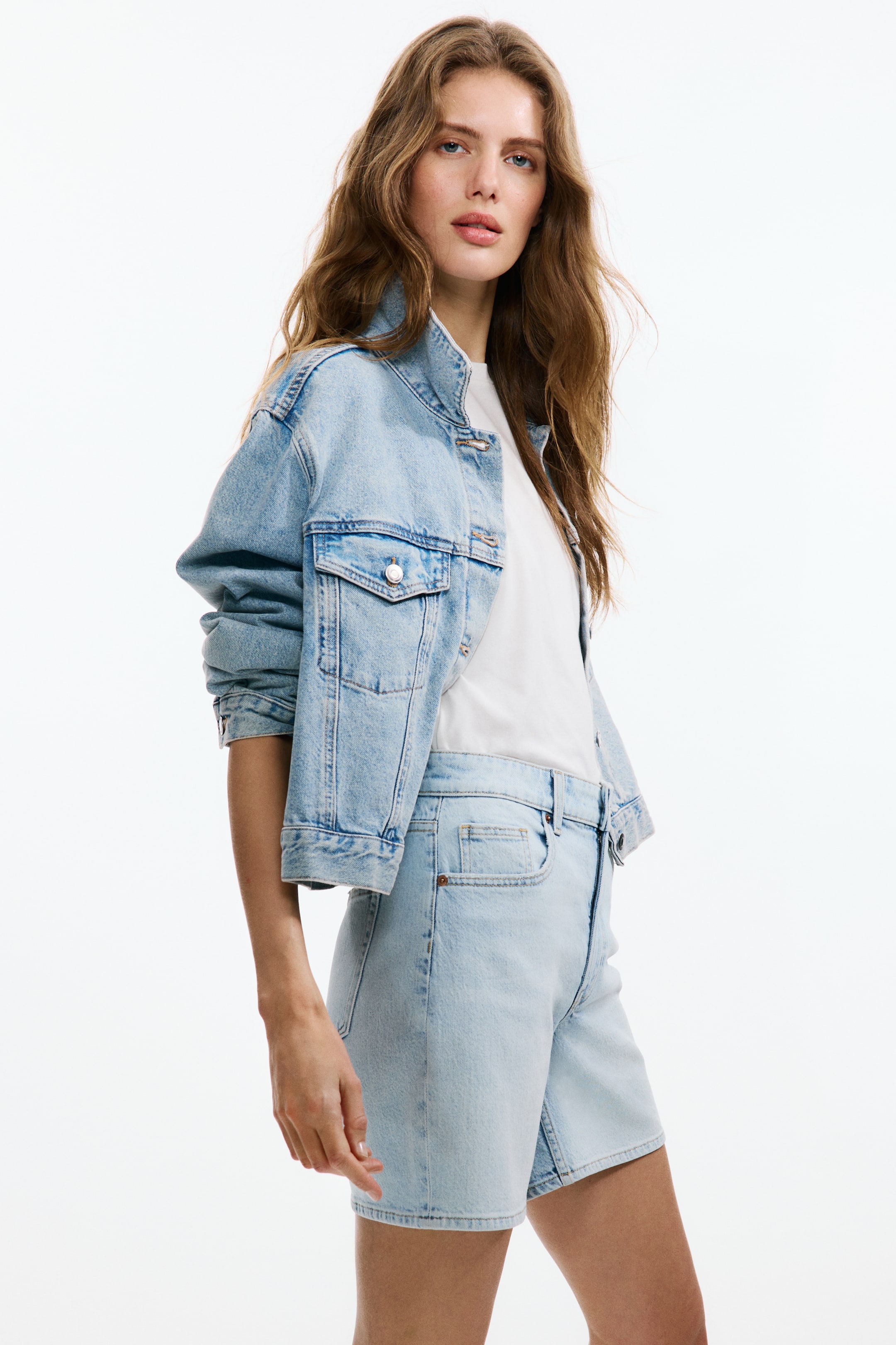 Visualizza immagine più grande: Una giacca di jeans azzurro chiaro è presentata, caratterizzata da un colletto alla coreana, spalle cadenti e tasche sul petto con patta e bottoni automatici, indossata sopra una t-shirt bianca. Sono indossati anche shorts di jeans azzurro chiaro coordinati, che presentano una vita alta e una vestibilità rilassata.