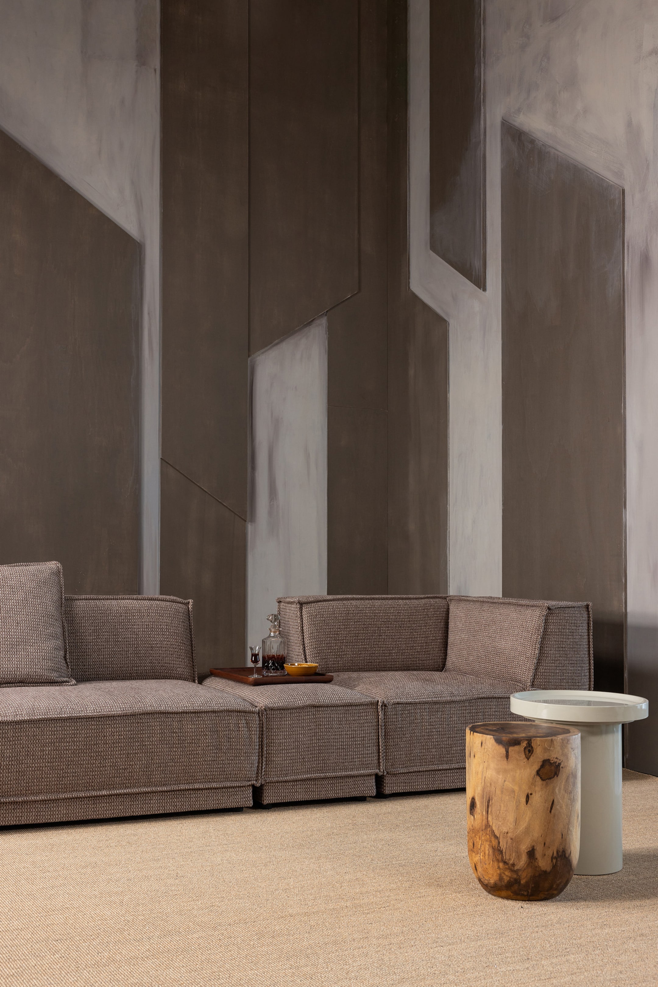 Agrandir l'image: Un canapé modulaire montre ses profils avant et latéral, recouvert d'un tissu brun texturé avec des coutures apparentes le long de ses bords. Un plateau en bois portant une carafe, deux verres, et un petit bol jaune repose sur l'un des modules du canapé.