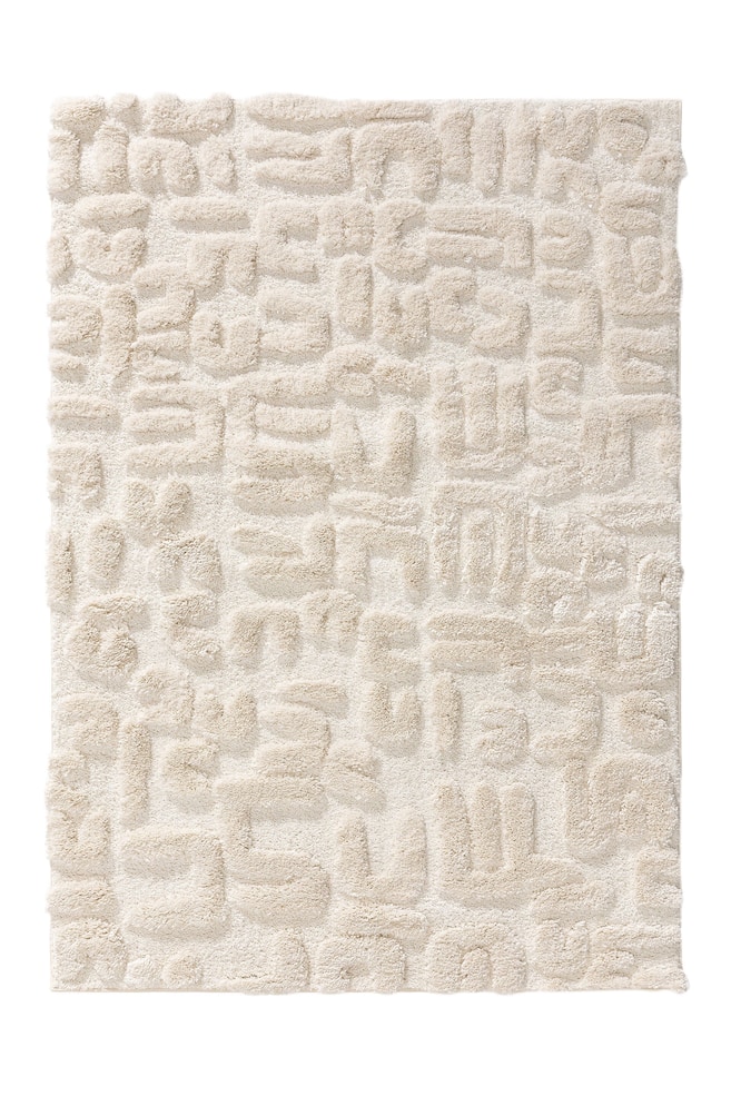 Un tapis rectangulaire de couleur crème présente une texture à poils longs. Il arbore un motif en relief répétitif de formes abstraites et massives dans une teinte de crème légèrement plus foncée, créant un effet de relief sur toute sa surface. - 1