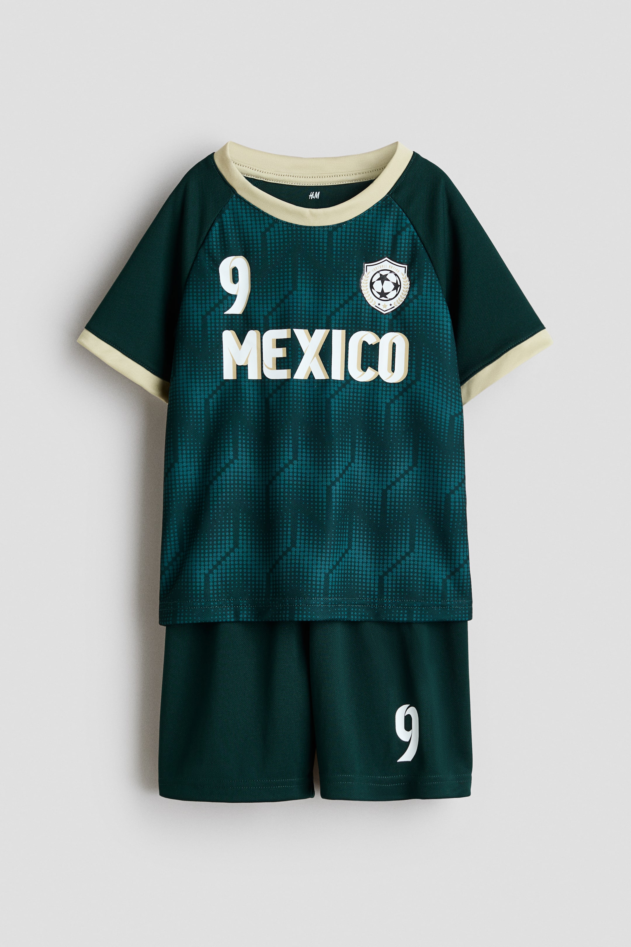 View larger image: A printed soccer set in soft mesh fabric is shown front on, featuring a dark green short-sleeved top with a geometric dot pattern in varying shades of green, light beige trim on the round neck and sleeve cuffs, a white number '9' on the left chest, 'MEXICO' printed below it, and a soccer emblem on the right chest. Matching solid dark green shorts, designed with an elasticated, drawstring waist, complete the set and feature a white number '9' on the lower right leg.