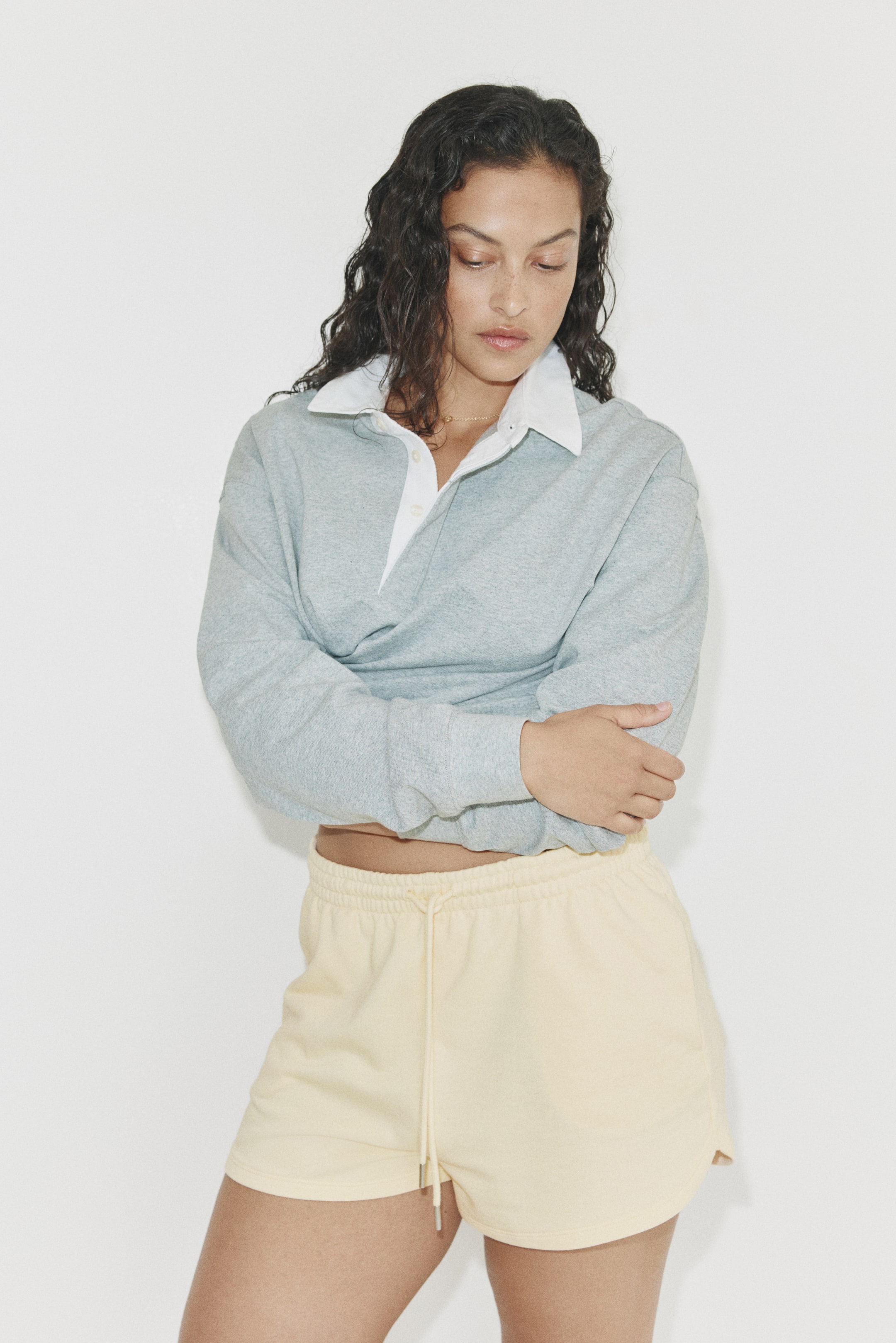View larger image: A light grey long-sleeved polo sweatshirt featuring a white collar and a partial button placket, with relaxed sleeves extending down. It's styled with light yellow shorts, which have an elastic waistband with a visible drawstring and curved leg openings.