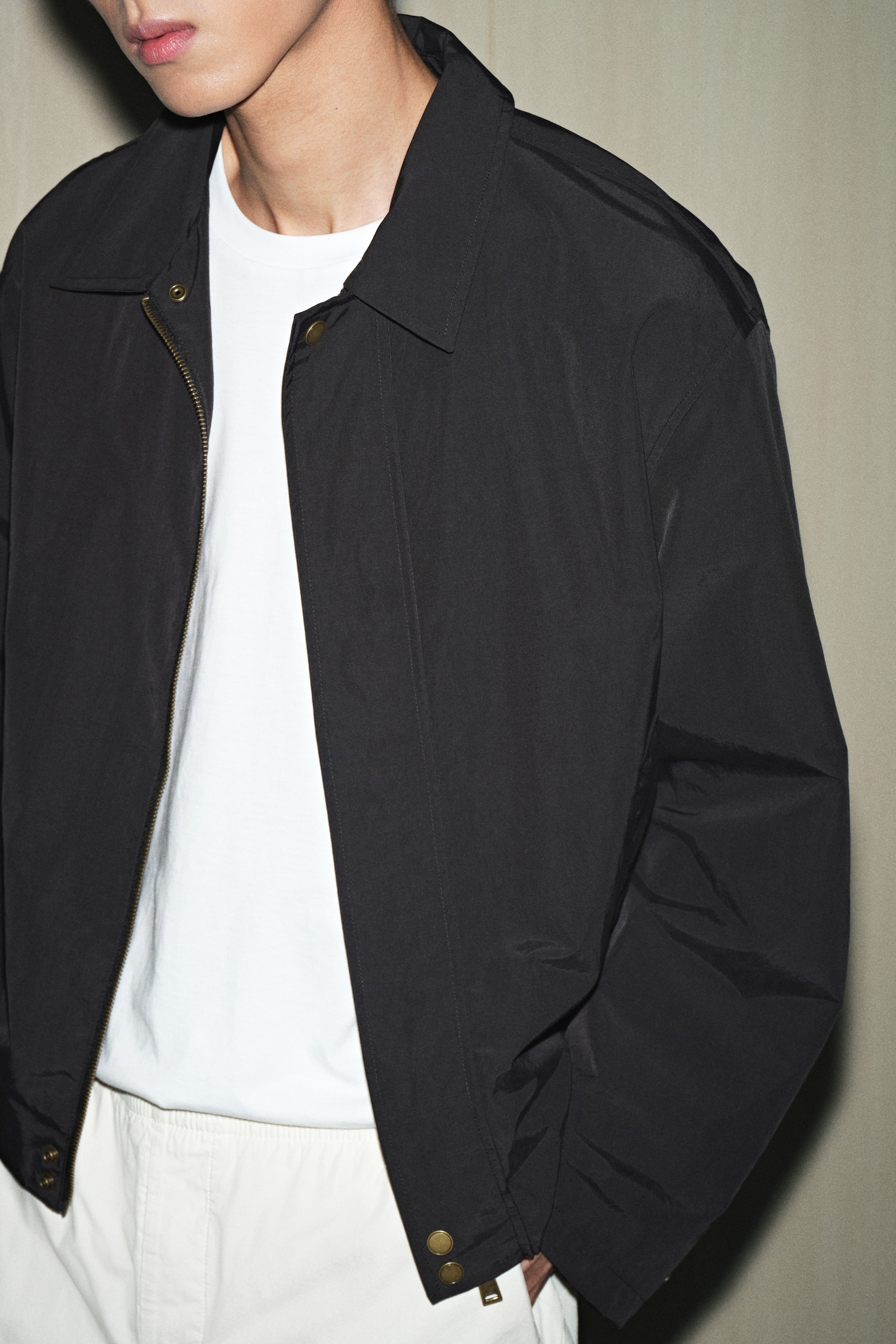 拡大画像を表示: 黒いジャケットが前面から、やや右に傾けて示されています。ポイントカラーと真鍮色のジッパーを備えたフルジップの前面閉鎖が特徴です。リラックスフィットのジャケットは、下部の裾に2つの真鍮色のスナップ留めがあり、その下には白いクルーネックのTシャツが着用されています。