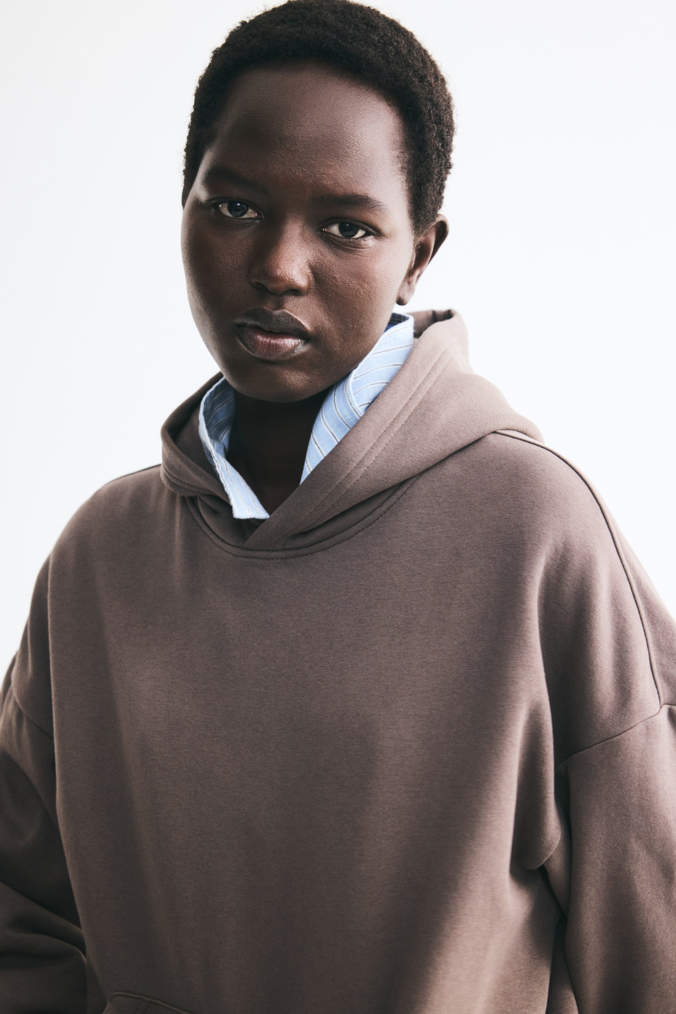 Visa större bild: En hoodie i taupebrun sweatshirtväv med dubbellagerhuva och sänkta axlar bärs av modellen. Hoodien är lagerlagd över en ljusblå randig skjortkrage som syns vid halsringningen.