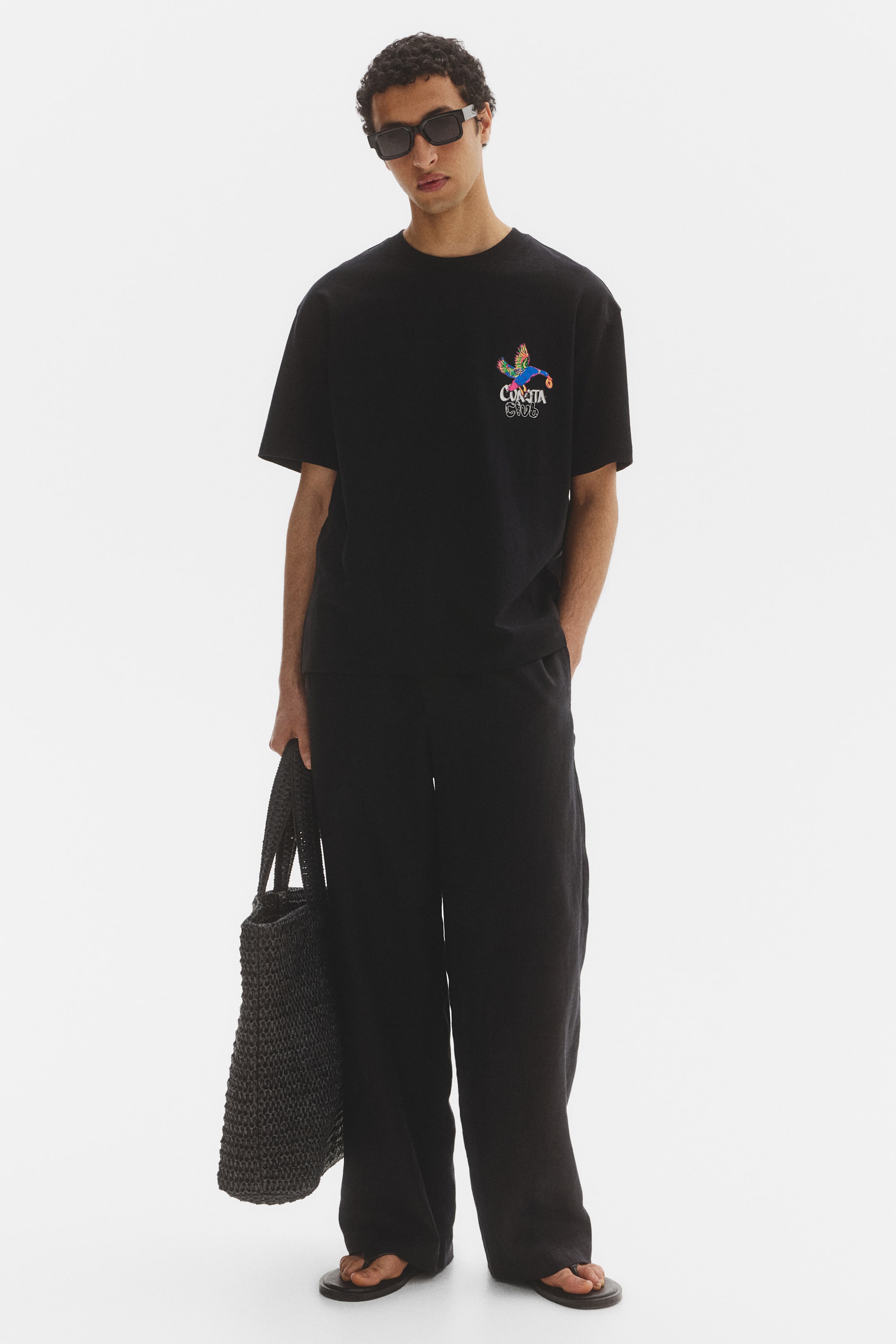 View larger image: An oversized black t-shirt showcases a vibrant graphic of a bird alongside the "CURTITA CLUB" text on the left chest. It's paired with black wide-leg trousers and black thong sandals. A black woven tote bag complements the look, while black rectangular sunglasses complete the ensemble.