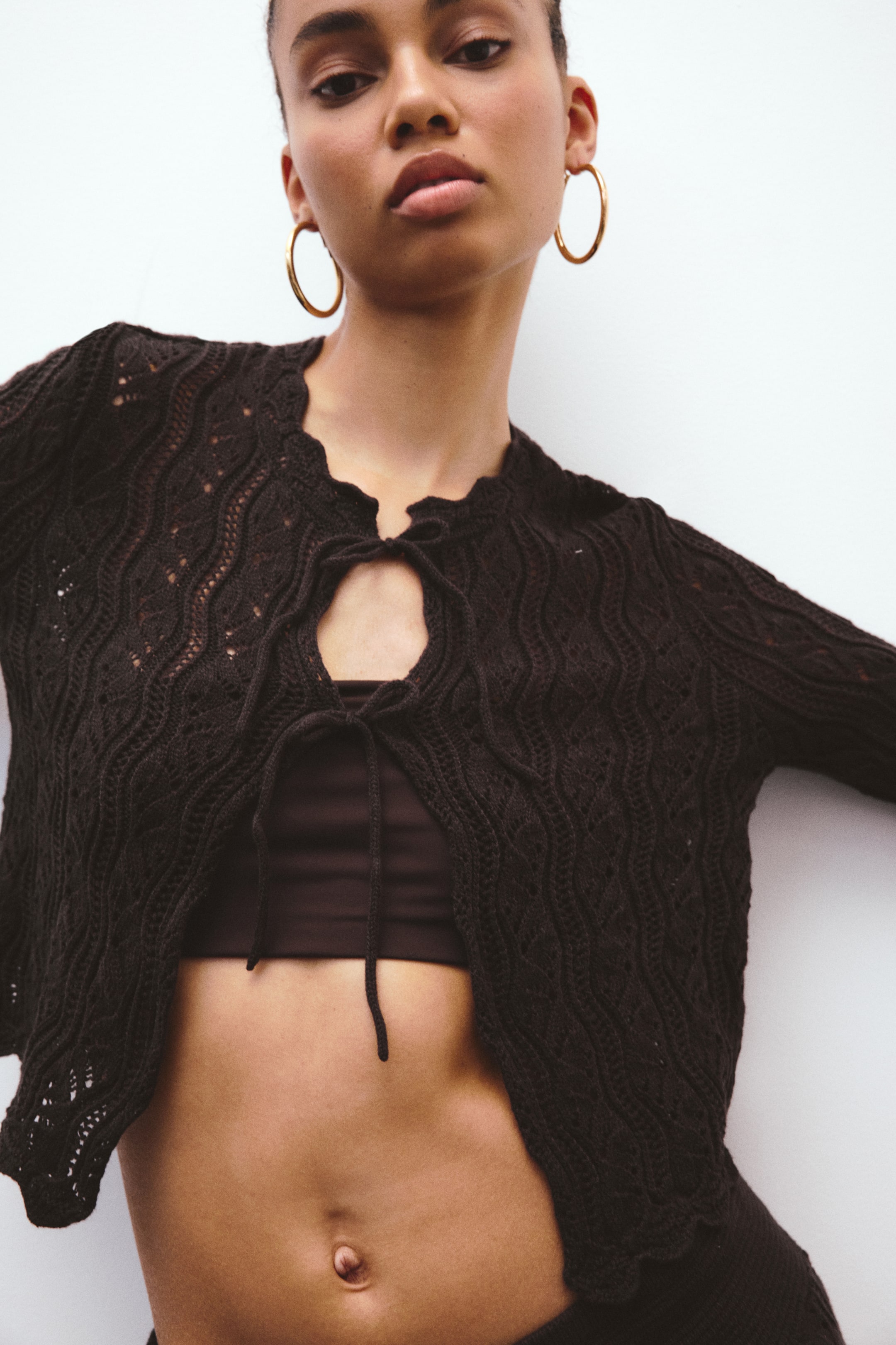 View larger image: The dark brown open-knit cardigan showcases a wavy design and scalloped edges, featuring a tie-front closure that creates a keyhole opening. It is styled over a dark brown bra top, highlighting its cropped fit.