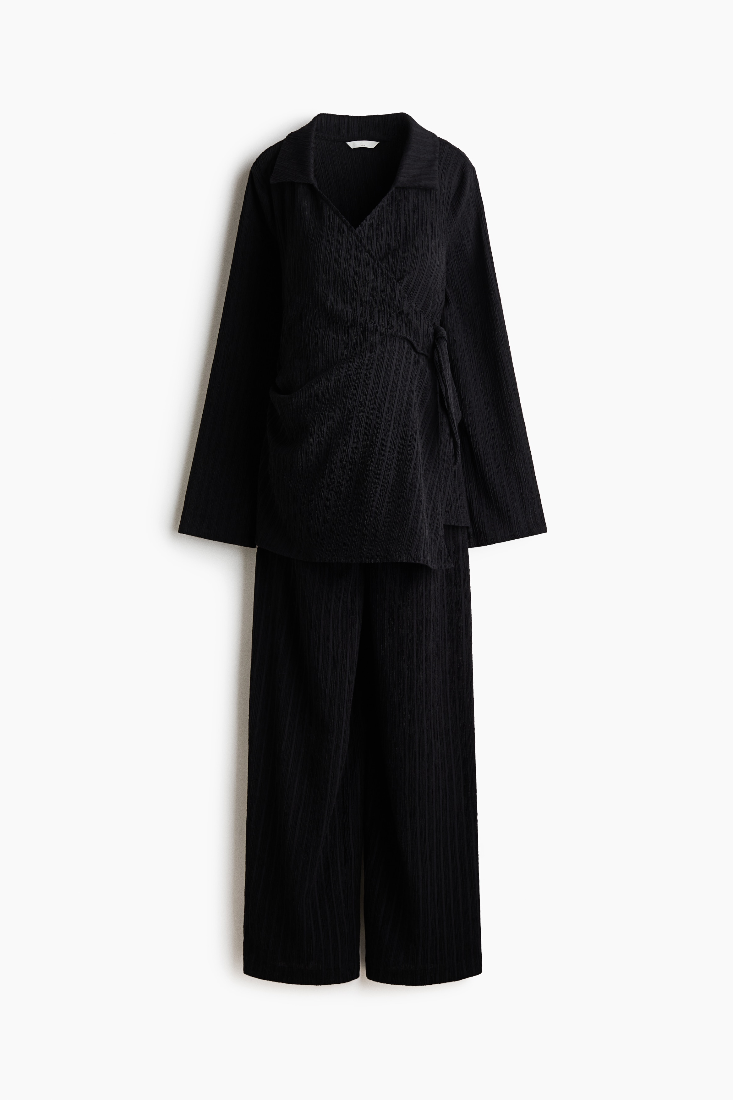 A black textured jersey set, featuring a long-sleeved wrap top with a collar and matching wide-leg trousers, is being worn by the model. The model is adjusting the tie fastening at the side of the wrap top.
