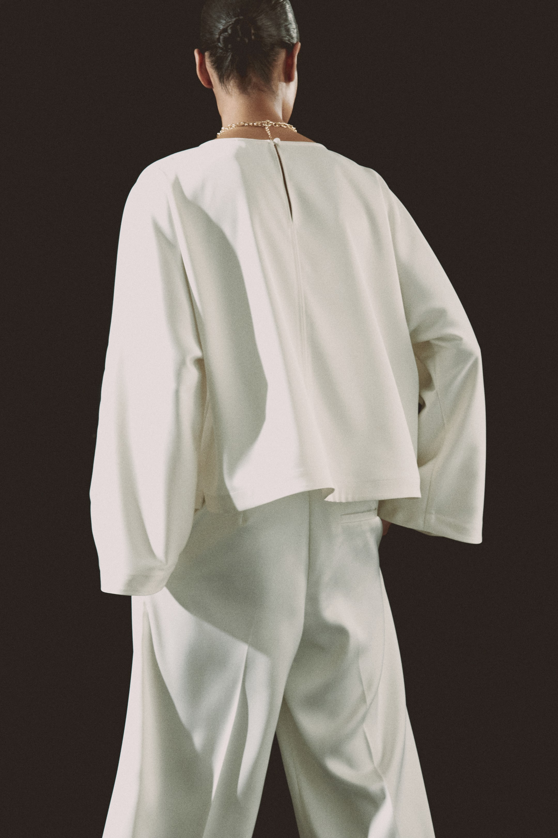 View larger image: A white, loose-fitting top is worn, showcasing wide, voluminous sleeves and a small keyhole opening with a button closure at the back of the neck. It is paired with matching white wide-leg trousers, and a gold chain necklace rests on the top's back neckline.