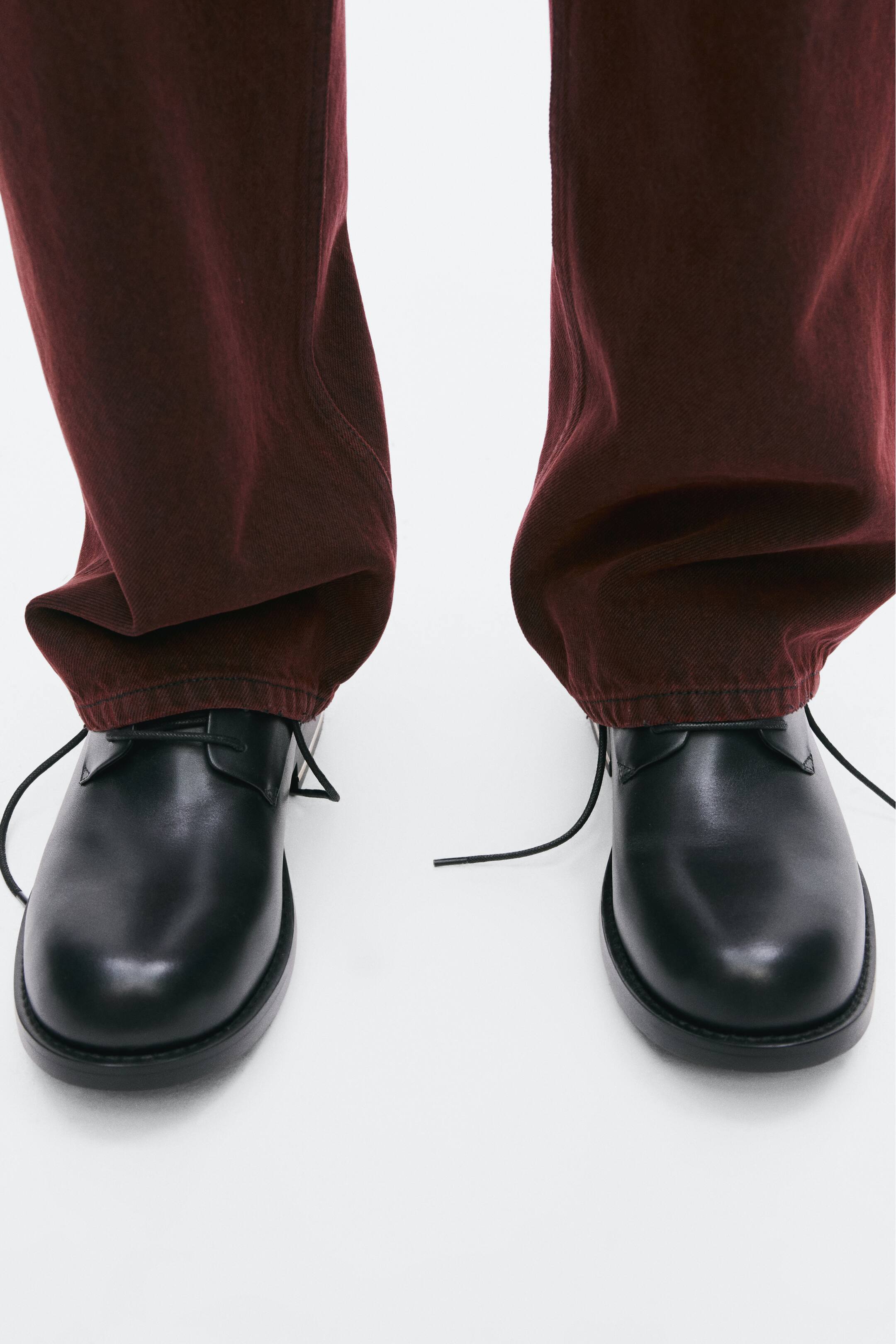 View larger image: The model is wearing black leather derby shoes, paired with dark burgundy trousers that fall slightly over the ankles. The shoes feature open lacing with thin, untied black laces and a rounded toe.