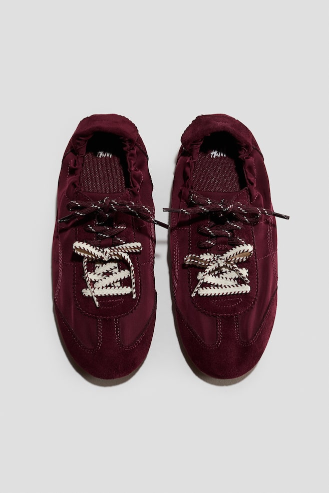 A pair of low-profile burgundy sneakers is shown from a top-down front view, featuring a combination of smooth textile and suede-textured panels. The sneakers are displaying an elasticated collar and a double-laced front with patterned laces in burgundy and cream. - 1