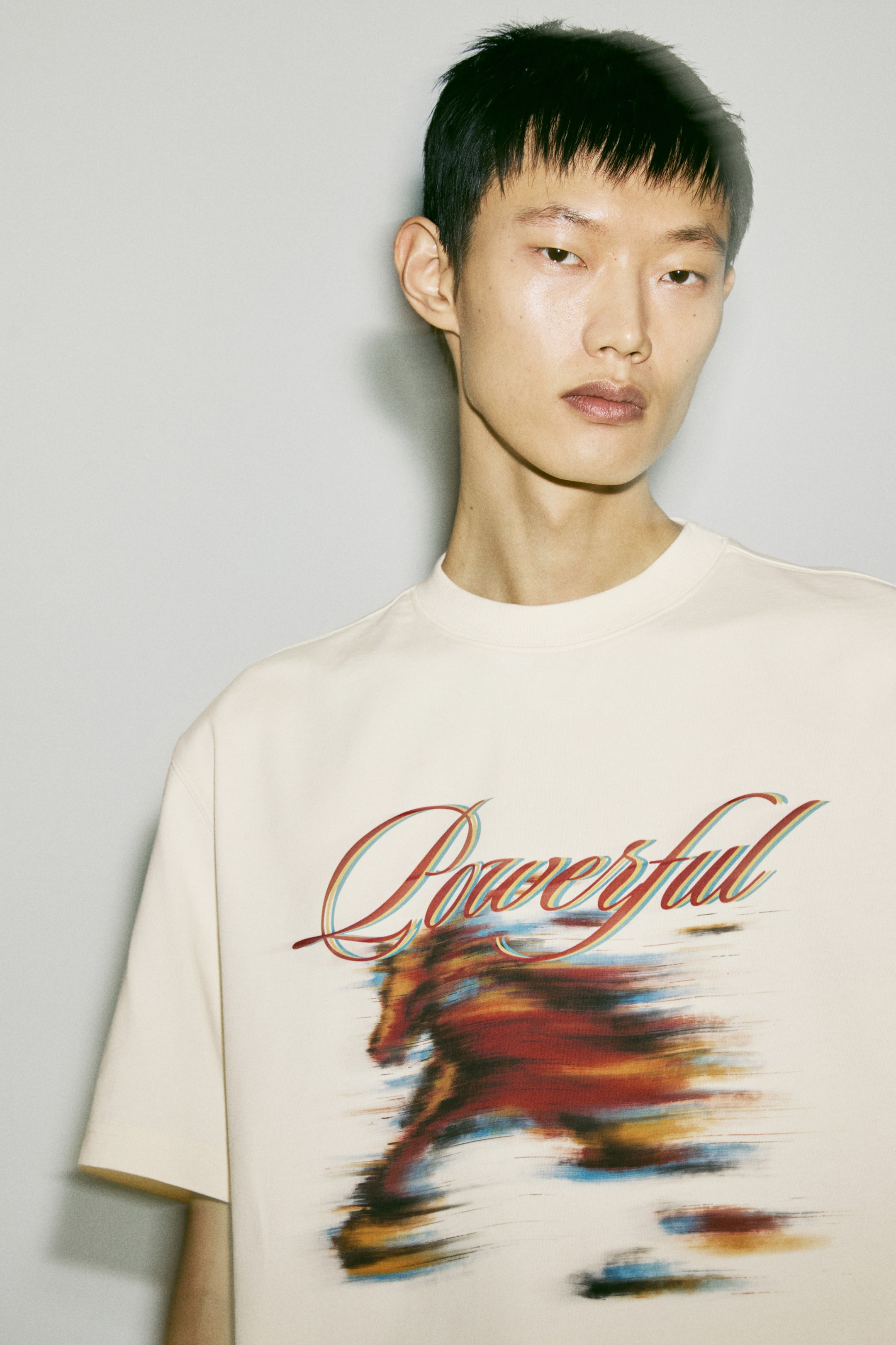Xem ảnh lớn hơn: Chiếc áo phông màu kem này có một họa tiết in lớn ở mặt trước. Họa tiết gồm từ "Powerful" được thể hiện bằng chữ viết tay cách điệu, xếp lớp màu đỏ và xanh dương, nằm trên một hình ảnh con ngựa có hiệu ứng mờ do chuyển động, được thể hiện qua các vệt động màu đỏ, cam, nâu và xanh dương. Áo có cổ tròn gân (rib) và tay ngắn.