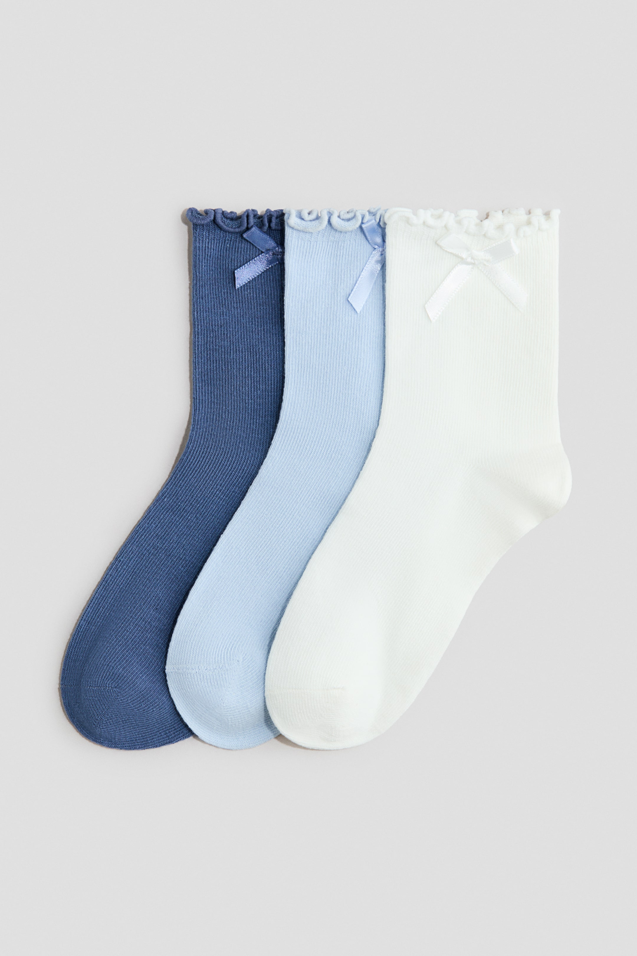Größeres Bild anzeigen: Drei Socken werden frontal präsentiert und zeigen ein feines, geripptes Strickmuster in Dunkelblau, Hellblau und Weiß. Jede Socke verfügt über einen elastischen, mit Rüschen besetzten Bund und eine kleine, passende Schleife, die die Vorderseite ziert.