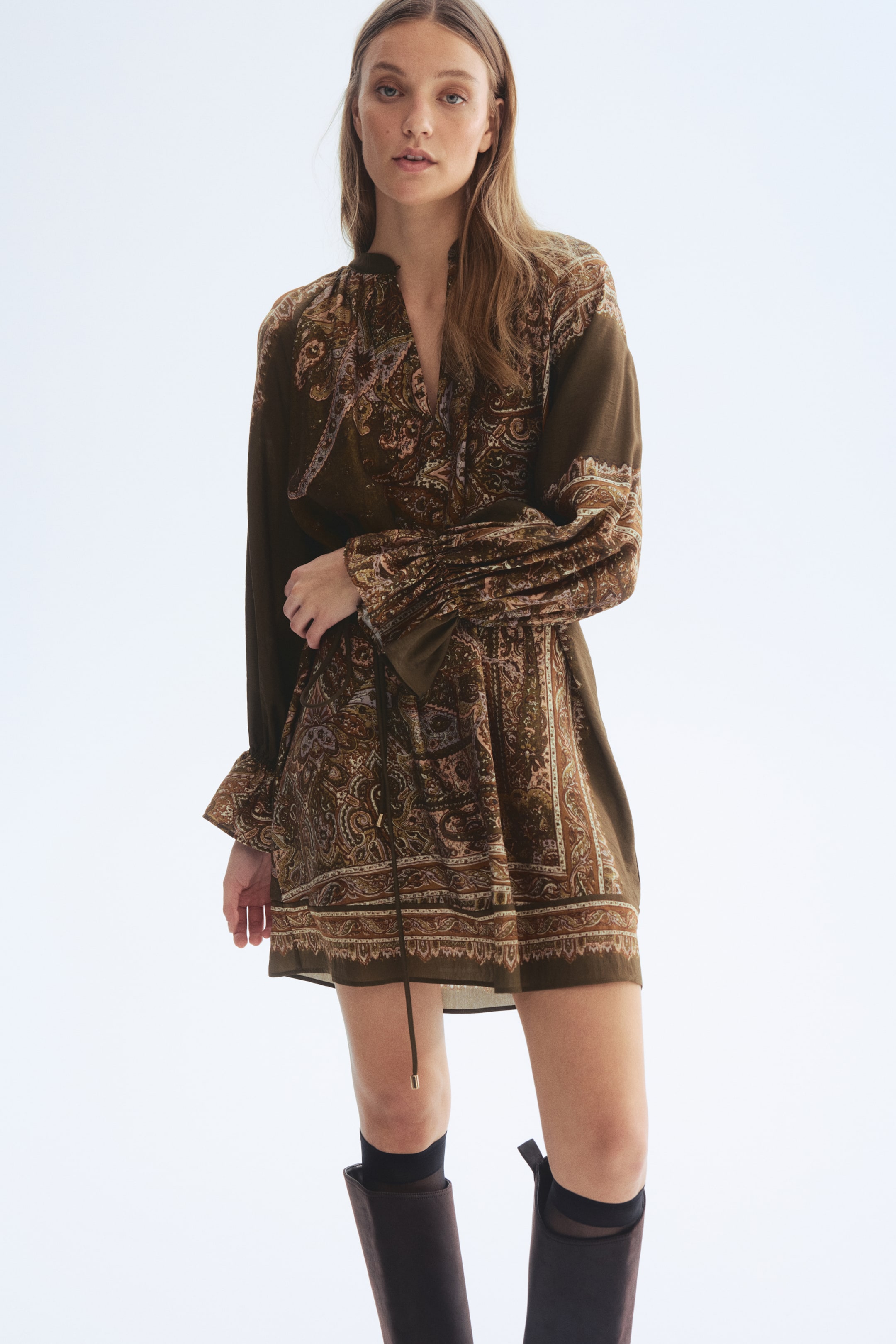 Zobrazit větší obrázek: Krátké šaty s olivově hnědým základem a složitým paisley vzorem, s výstřihem do V a stojatým límcem. Dlouhé, objemné rukávy končí nařasenými, volánkovými manžetami a šňůrka v pase vytváří splývavou siluetu. Šaty jsou prezentovány z mírného úhlu zepředu zleva.