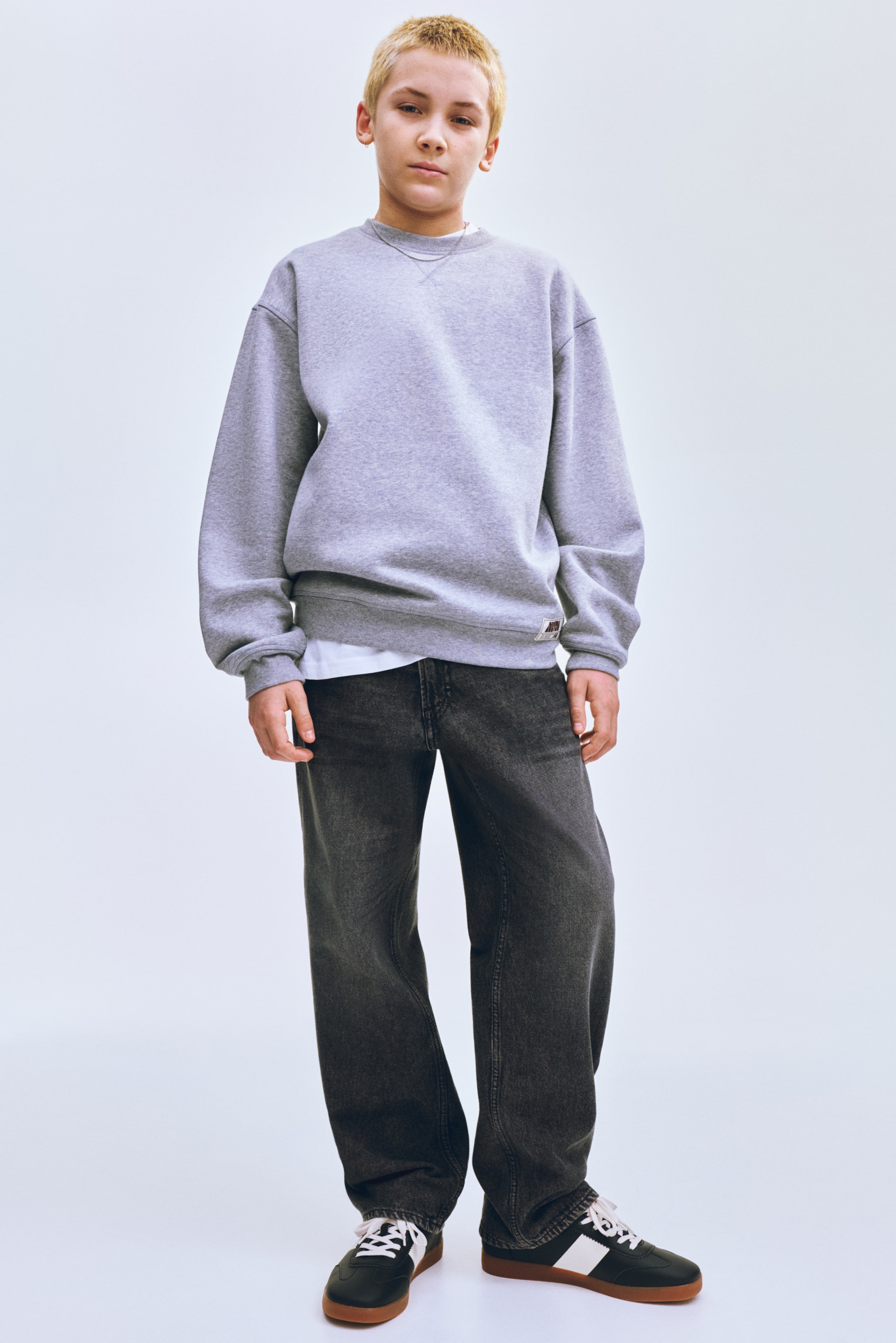 Grösseres Bild ansehen: Ein graues Rundhals-Sweatshirt wird präsentiert, das einen gerippten Ausschnitt mit V-Naht-Details, lange Ärmel mit gerippten Bündchen und einen gerippten Saum aufweist. Ein kleines rechteckiges Etikett ist auf der unteren linken Seite sichtbar, und ein weißes T-Shirt wird darunter getragen.