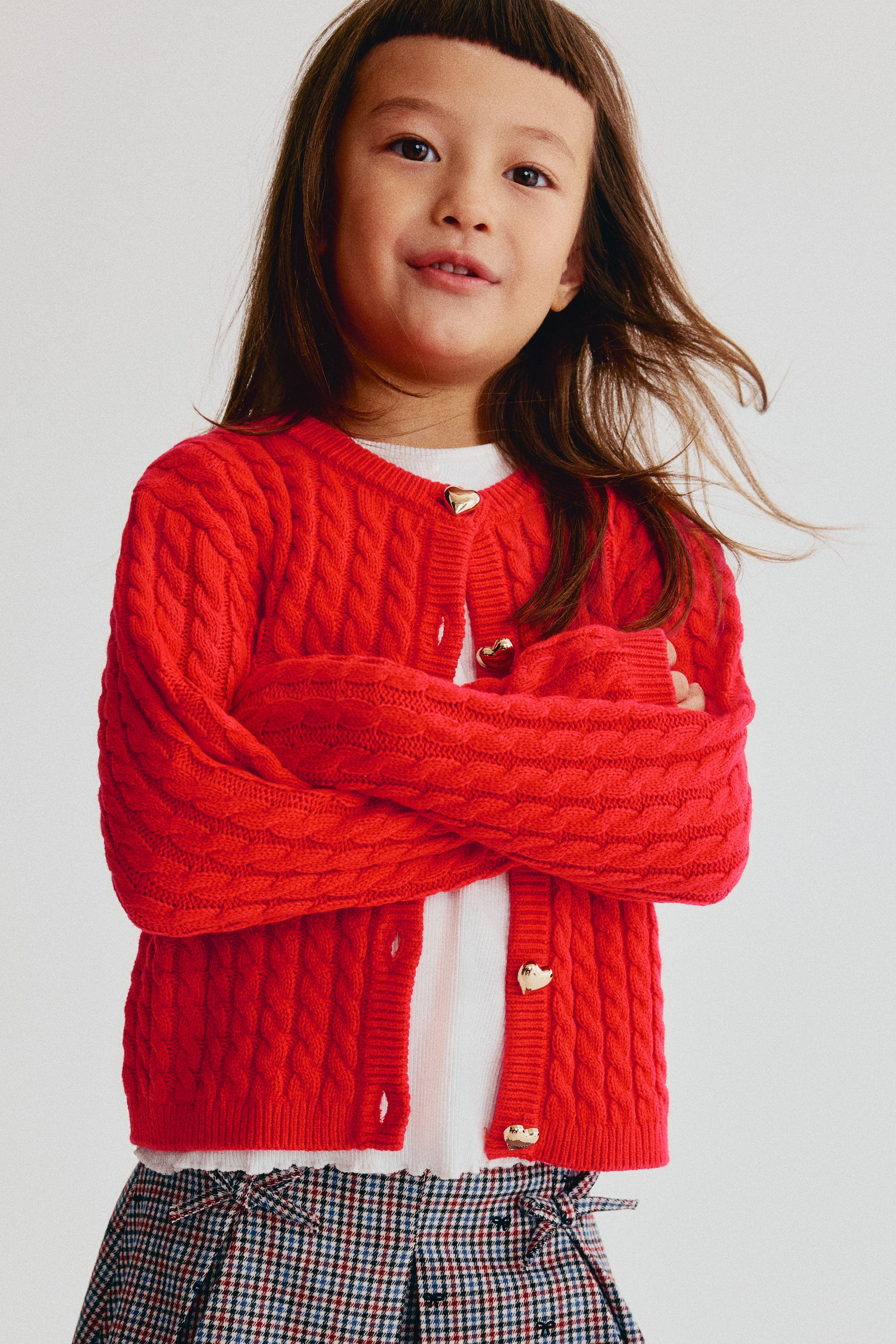 View larger image: A red cable-knit cardigan is shown from the front, featuring a round neckline, ribbed trims, and gold heart-shaped buttons down the button band. The cardigan is worn layered over a white top, displaying its textured knit pattern and ribbed cuffs.