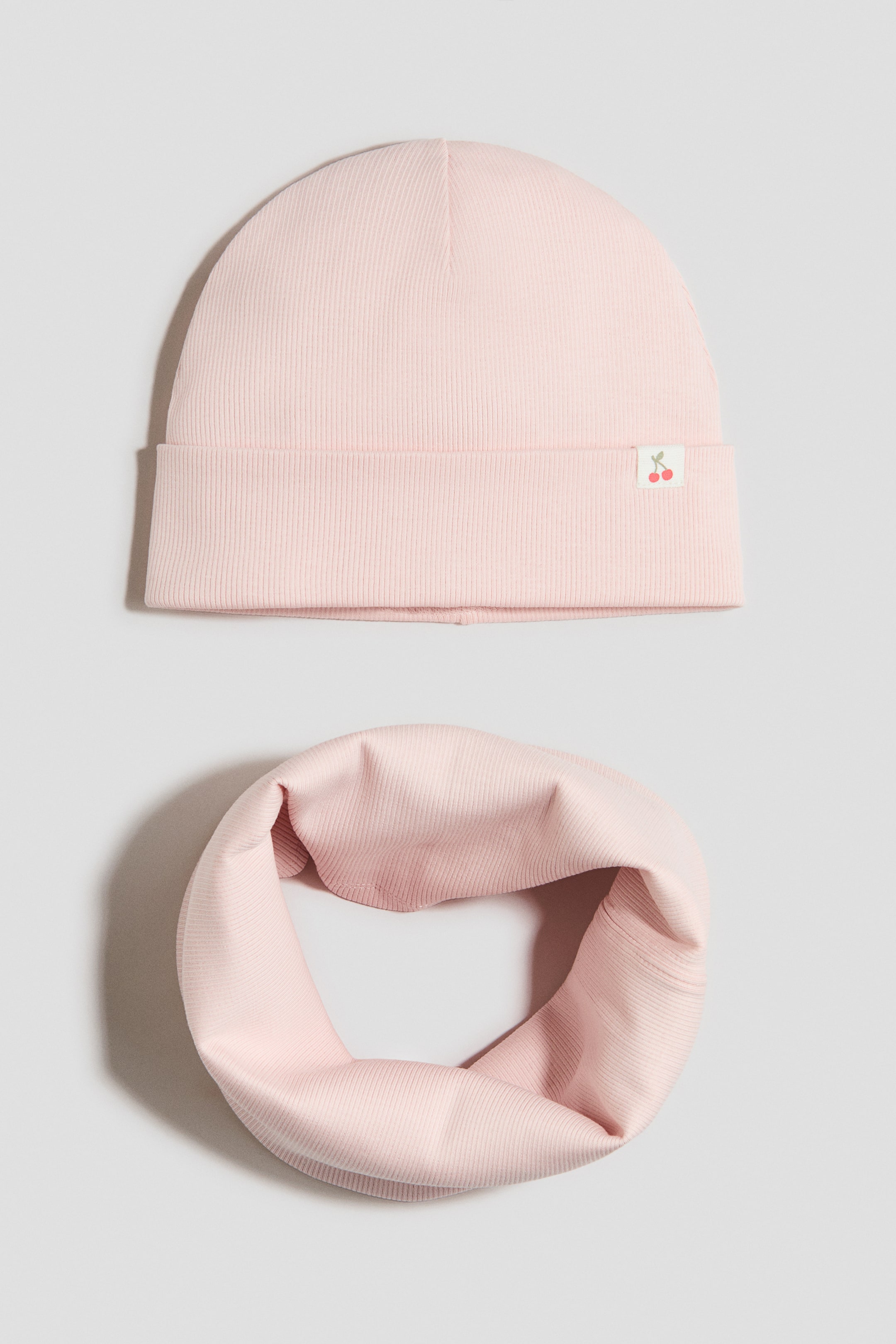 View larger image: A 2-piece jersey set displays a light pink ribbed beanie with a turn-up brim and a small white label bearing a red cherry motif on its front. A matching light pink ribbed neck warmer is presented as a circular, folded loop.