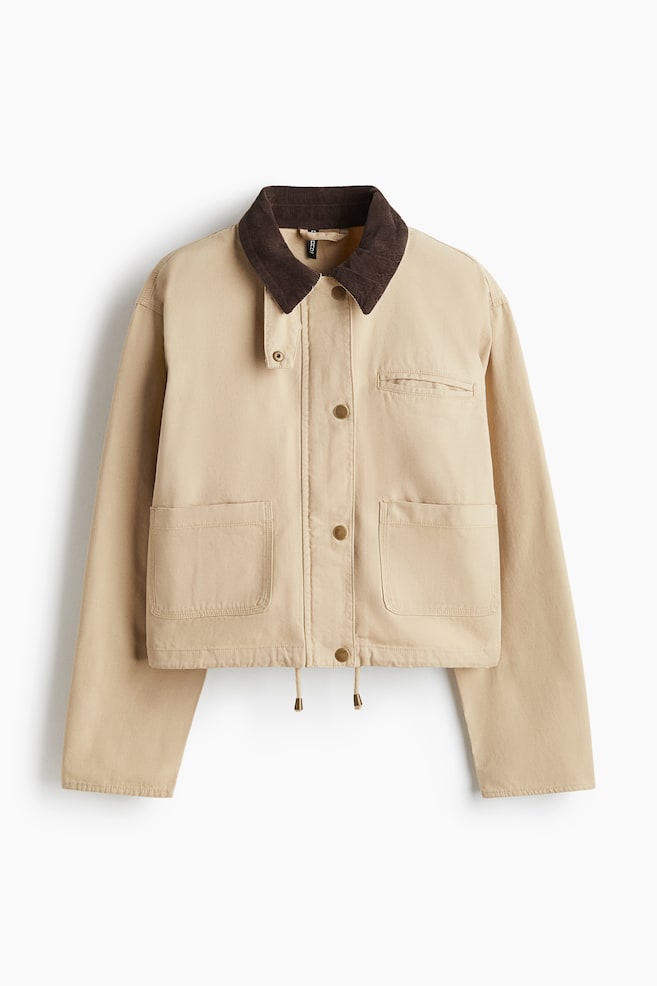 Eine nach vorne gerichtete, beige Jacke verfügt über einen dunkelbraunen gerippten Cordkragen mit einem Druckknopfverschluss auf der rechten Seite. Sie ist mit einem zentralen Knopfverschluss mit runden, hellbraunen Knöpfen, zwei großen aufgesetzten Taschen auf der unteren Vorderseite, einer kleinen horizontalen Schlitztasche auf der oberen linken Brust und einem Kordelzugsaum mit metallischen Kordelstoppern gestaltet. - 1