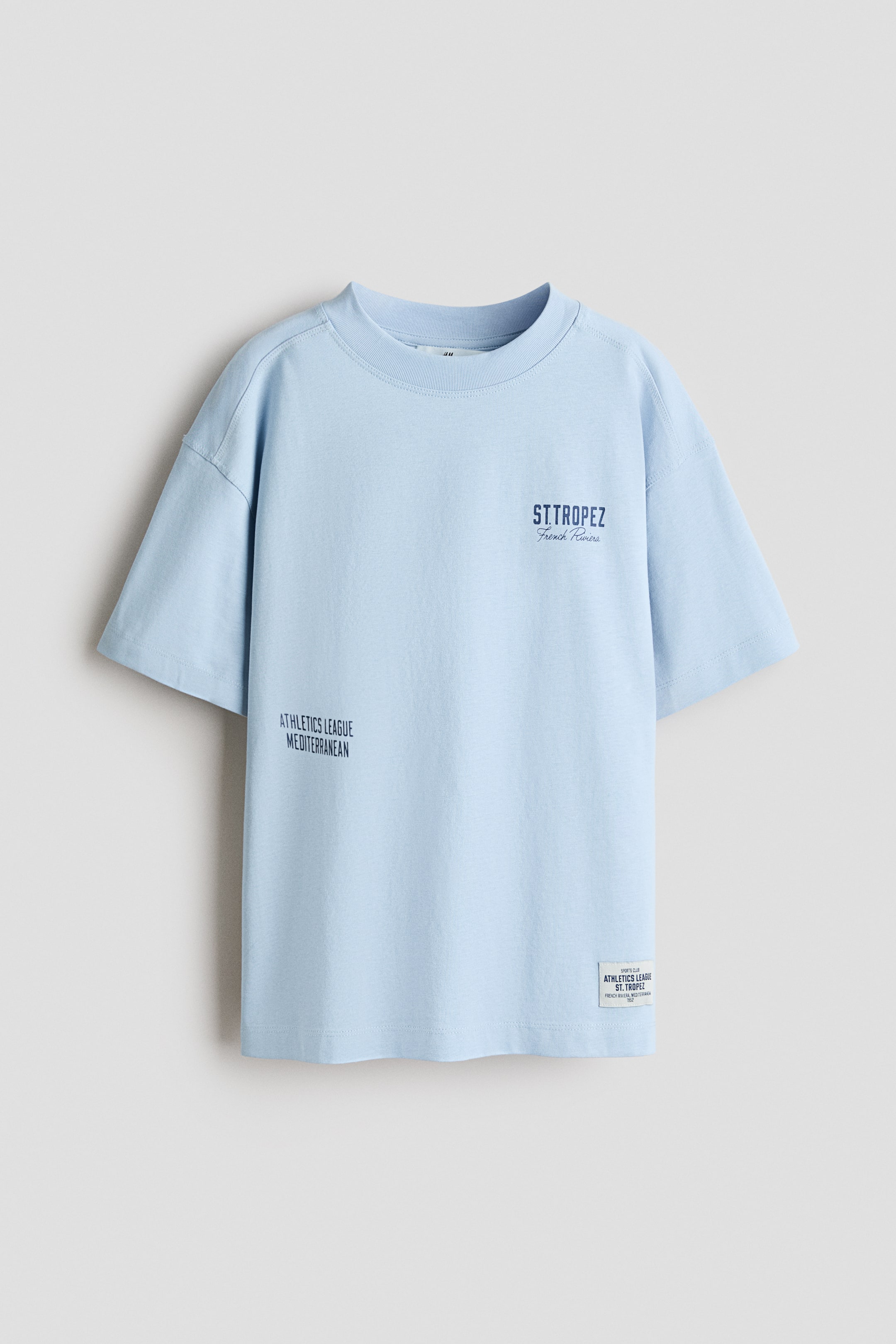 Grotere afbeelding bekijken: Een lichtblauw t-shirt wordt frontaal gepresenteerd, met een geribbelde ronde hals en korte, wijde mouwen met verlaagde schouders. Op de voorkant is donkerblauwe zeefdruktekst te zien, waaronder "ST.TROPEZ French Riviera" op de borst, "ATHLETICS LEAGUE MEDITERRANEAN" linksonder, en een klein wit stoffen label met donkere tekst rechtsonder.