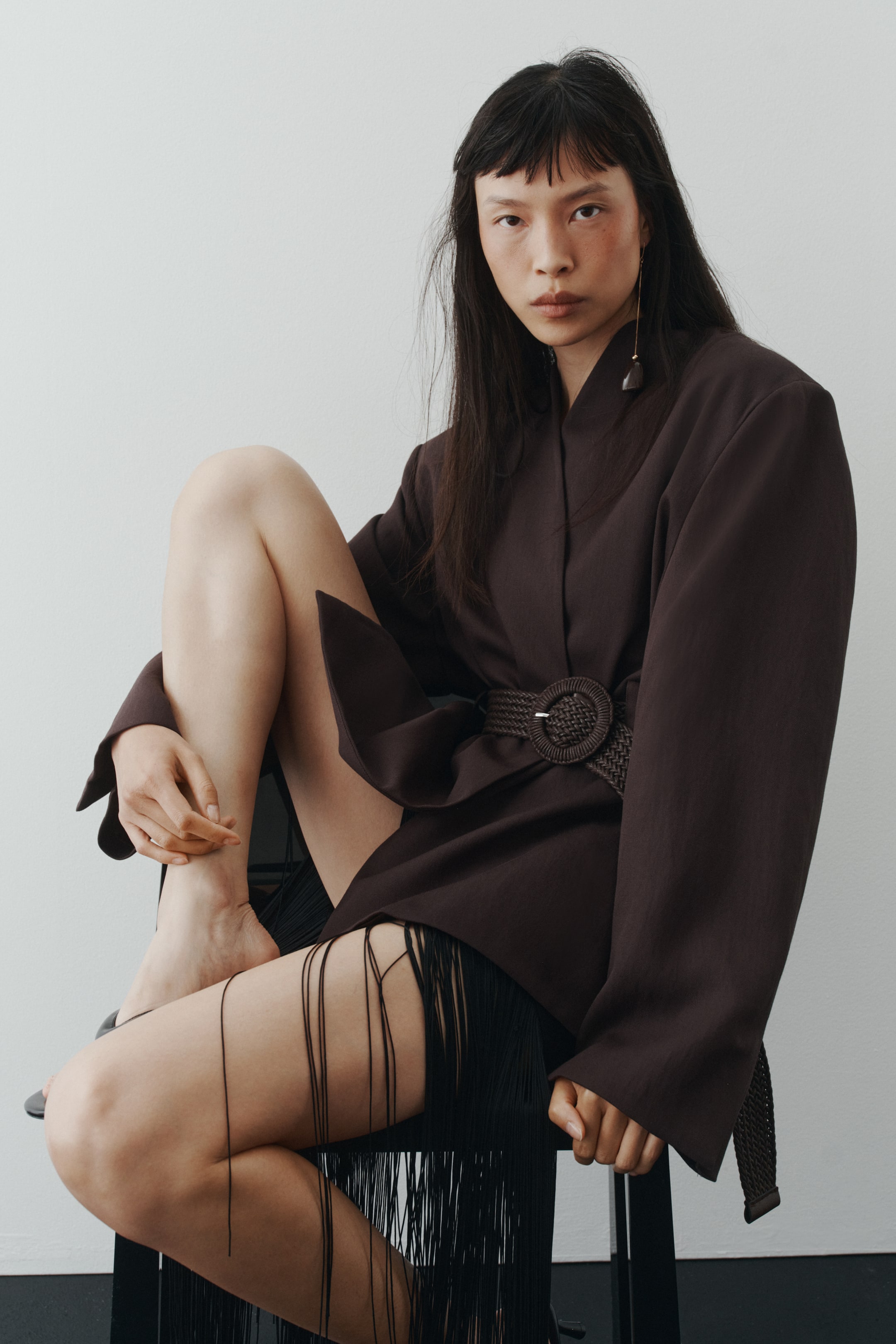 View larger image: The model is wearing a dark single-breasted twill blazer featuring a lapelless neckline, shoulder pads, and long sleeves with cuff slits. Its open front is elegantly cinched by a wide, braided waist belt with a round buckle. Beneath the blazer, a fringed garment is subtly visible.