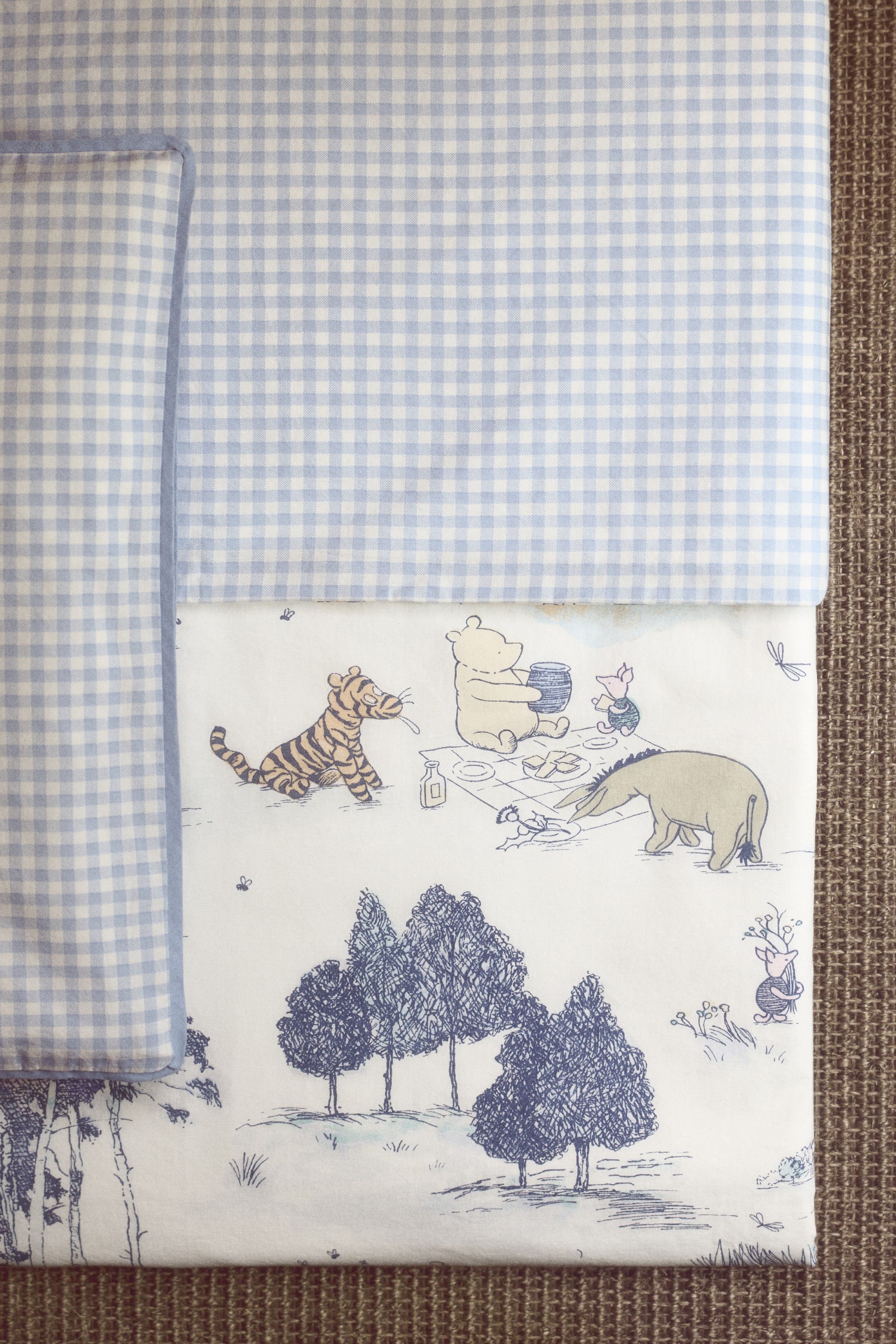 Agrandir l'image: Un ensemble de literie présente un motif à carreaux vichy bleu clair et blanc sur la partie supérieure, qui se replie sur un tissu blanc affichant des illustrations esquissées des personnages de Winnie l'Ourson pique-niquant parmi les arbres.