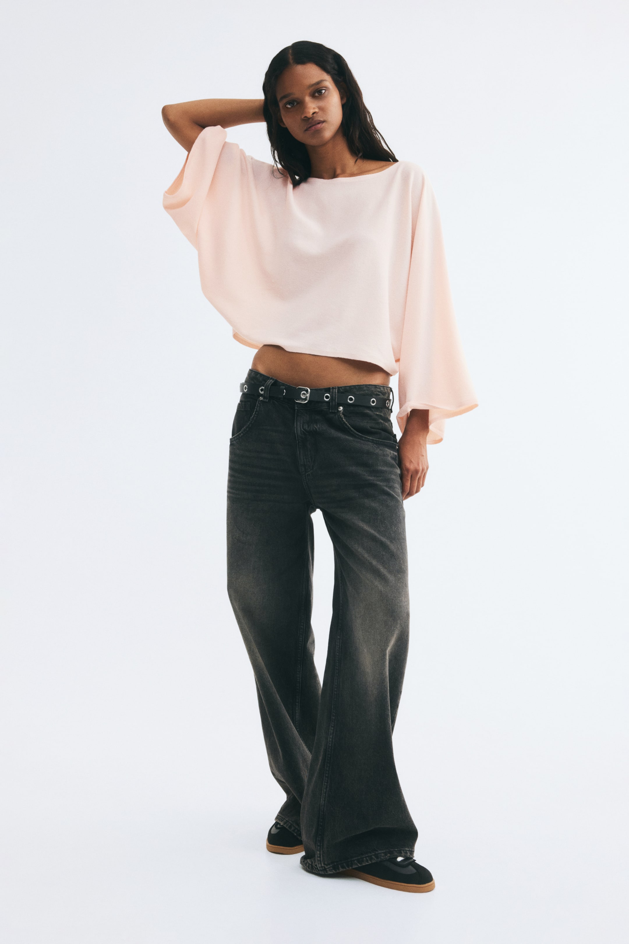 Voir une image plus grande: Un haut court à coupe ample, rose pâle, avec encolure bateau et longues manches cape, est porté par le mannequin, qui pose avec une main derrière la tête pour mettre en valeur le design drapé des manches. Le haut est agencé avec un jeans gris foncé à jambe large et une ceinture noire à œillets.