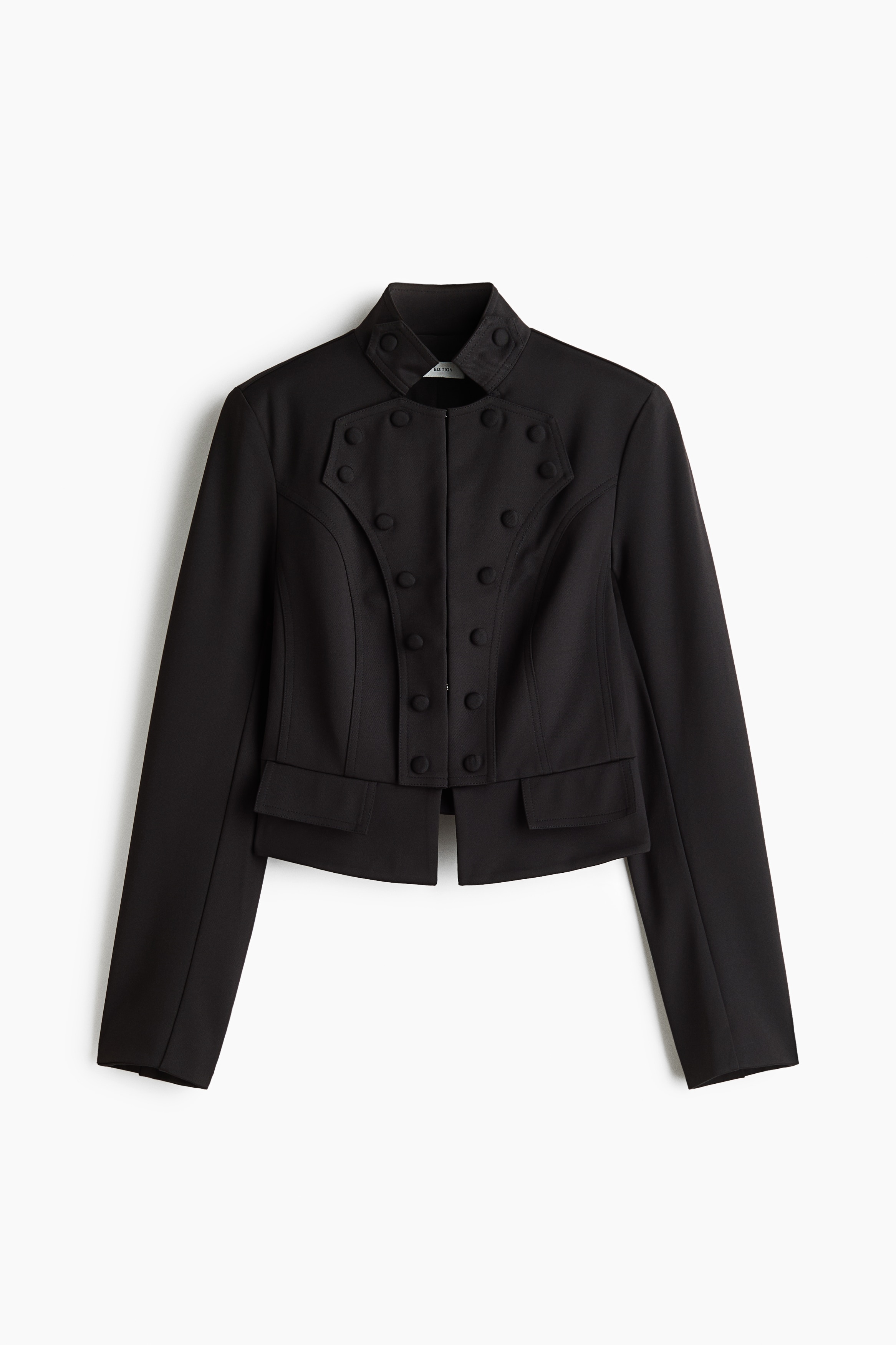 Una chaqueta corta negra con cuello mao, botones forrados decorativos en el delantero y tapetas de bolsillo frontales, que luce la modelo. La chaqueta se lleva abierta sobre una blusa blanca con cuello alto y volantes, y se combina con pantalones negros.