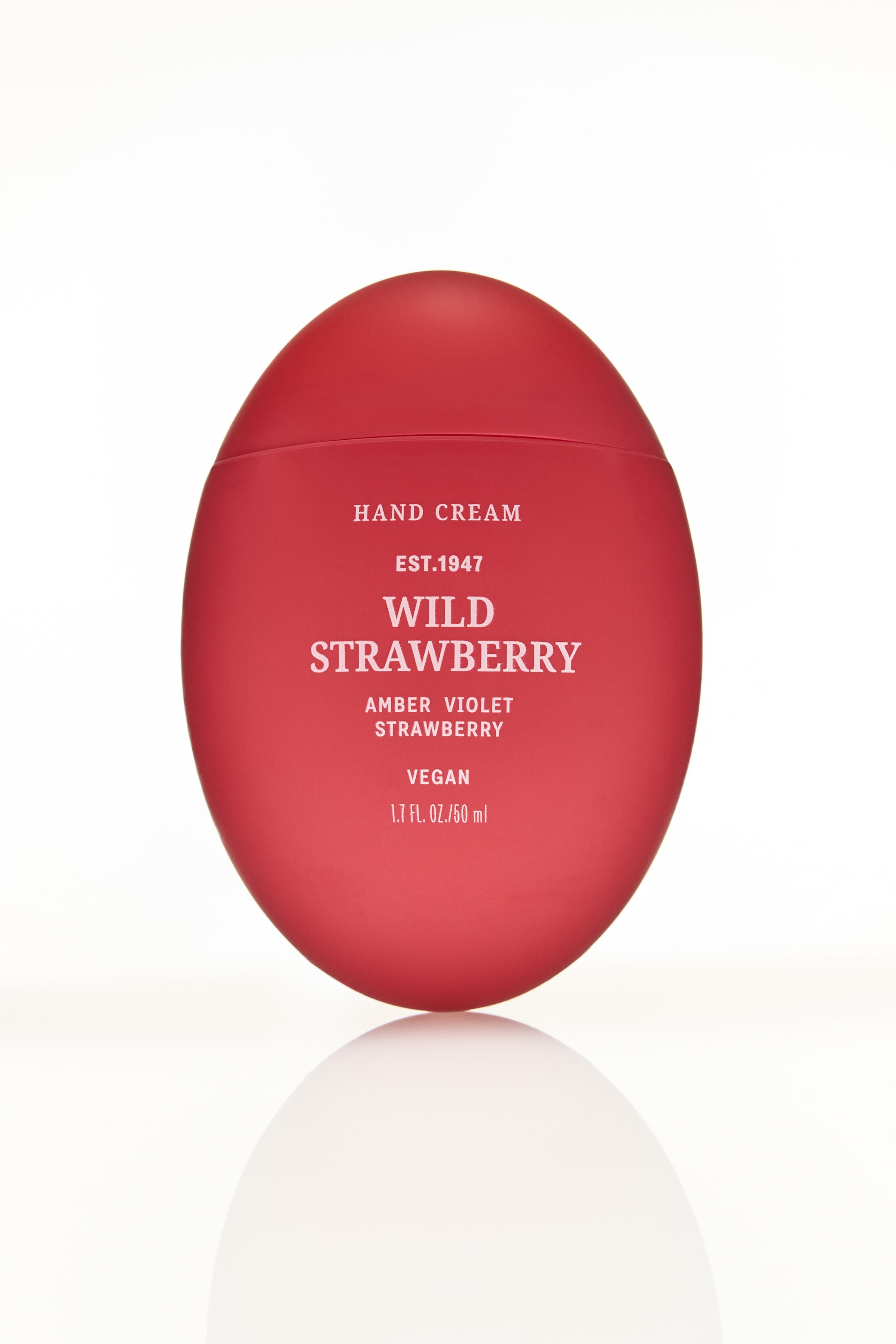 Ampliar la imagen: Un envase de crema de manos de color rojo mate y forma ovalada está orientado hacia adelante, mostrando texto en blanco que incluye "CREMA DE MANOS" y "FRESA SILVESTRE", con una costura horizontal visible cerca de la parte superior.