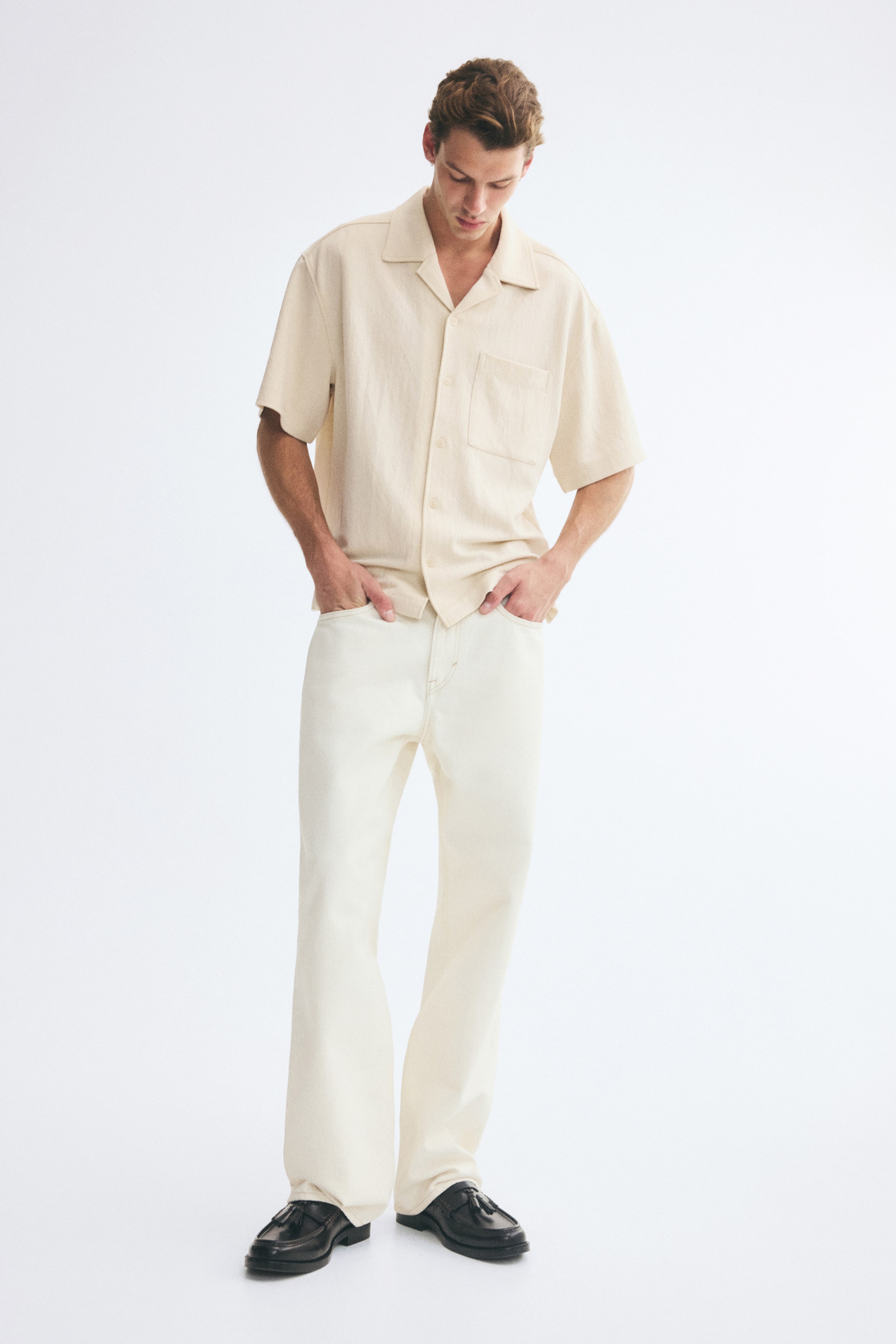View larger image: The model is wearing a light beige, short-sleeve jersey shirt with a resort collar, French front, and a single patch chest pocket. The relaxed-fit shirt is styled untucked over matching off-white trousers, with the model’s hands resting in the pockets.