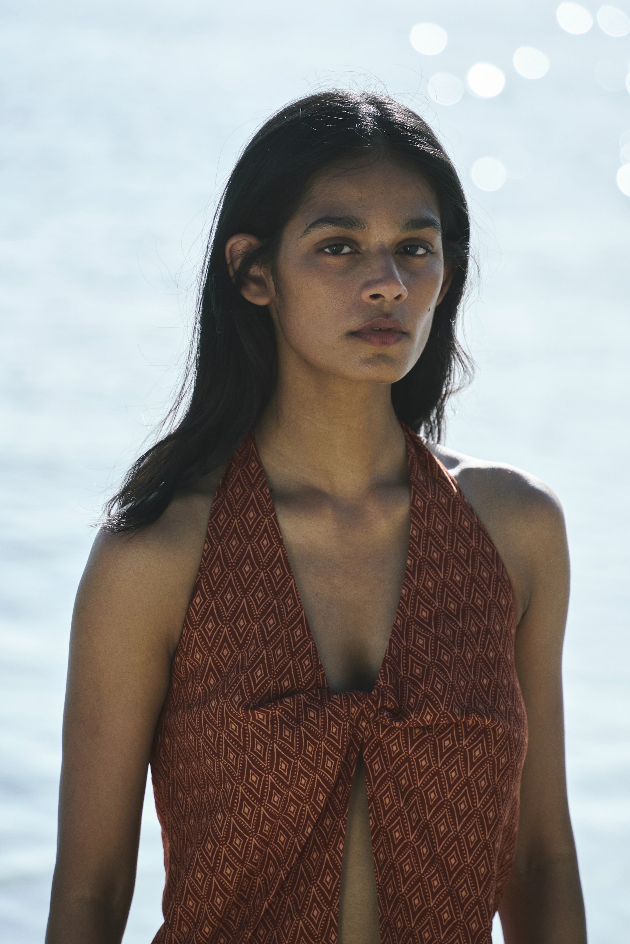View larger image: A halter neck top featuring a reddish-brown fabric with a geometric diamond pattern. It showcases a deep V-neckline and a knotted detail at the center front, forming a split opening.