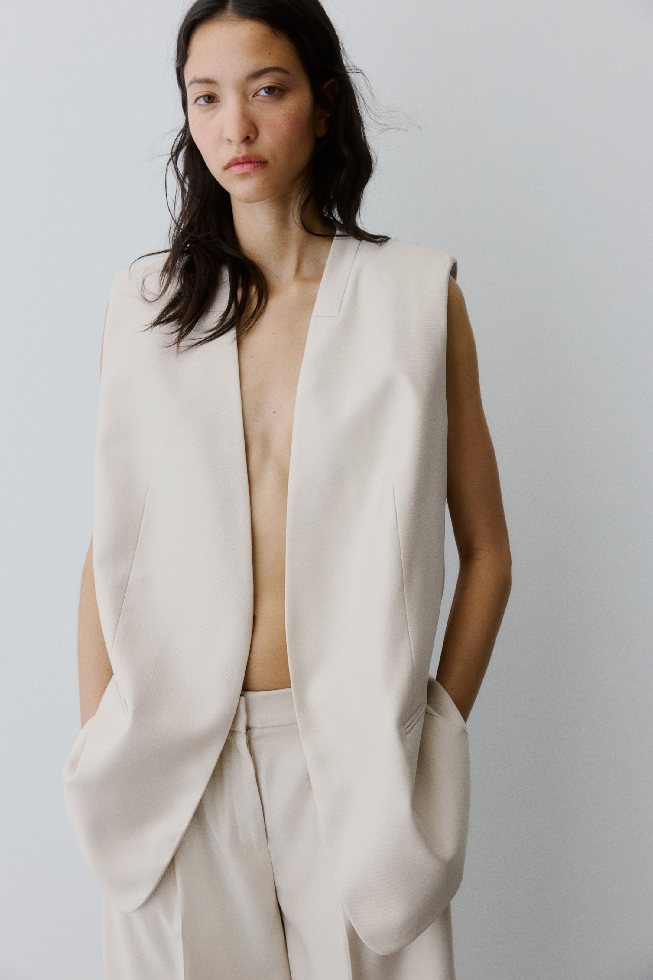 Agrandir l'image: Un long gilet tissé de couleur claire avec des épaules structurées et des poches passepoilées à l'avant est porté par le mannequin. Le gilet est ouvert, et le mannequin repose ses mains dans ses poches, complétant le look avec un pantalon assorti.