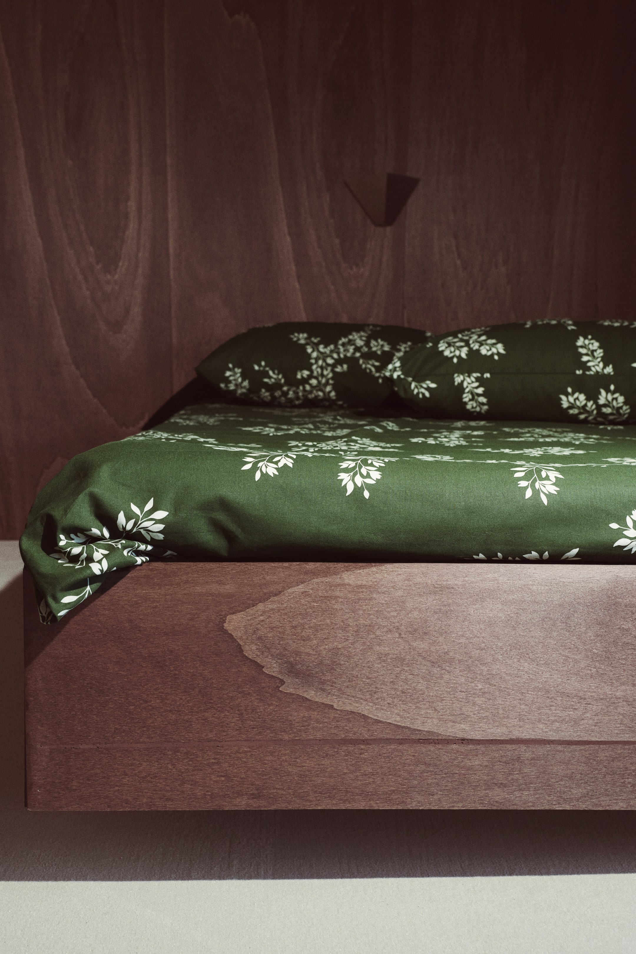 View larger image: A dark green cotton bedsheet set featuring an all-over white leaf-and-branch pattern is dressing a bed, with the flat sheet draping over the corner and two matching pillowcases resting at the head.