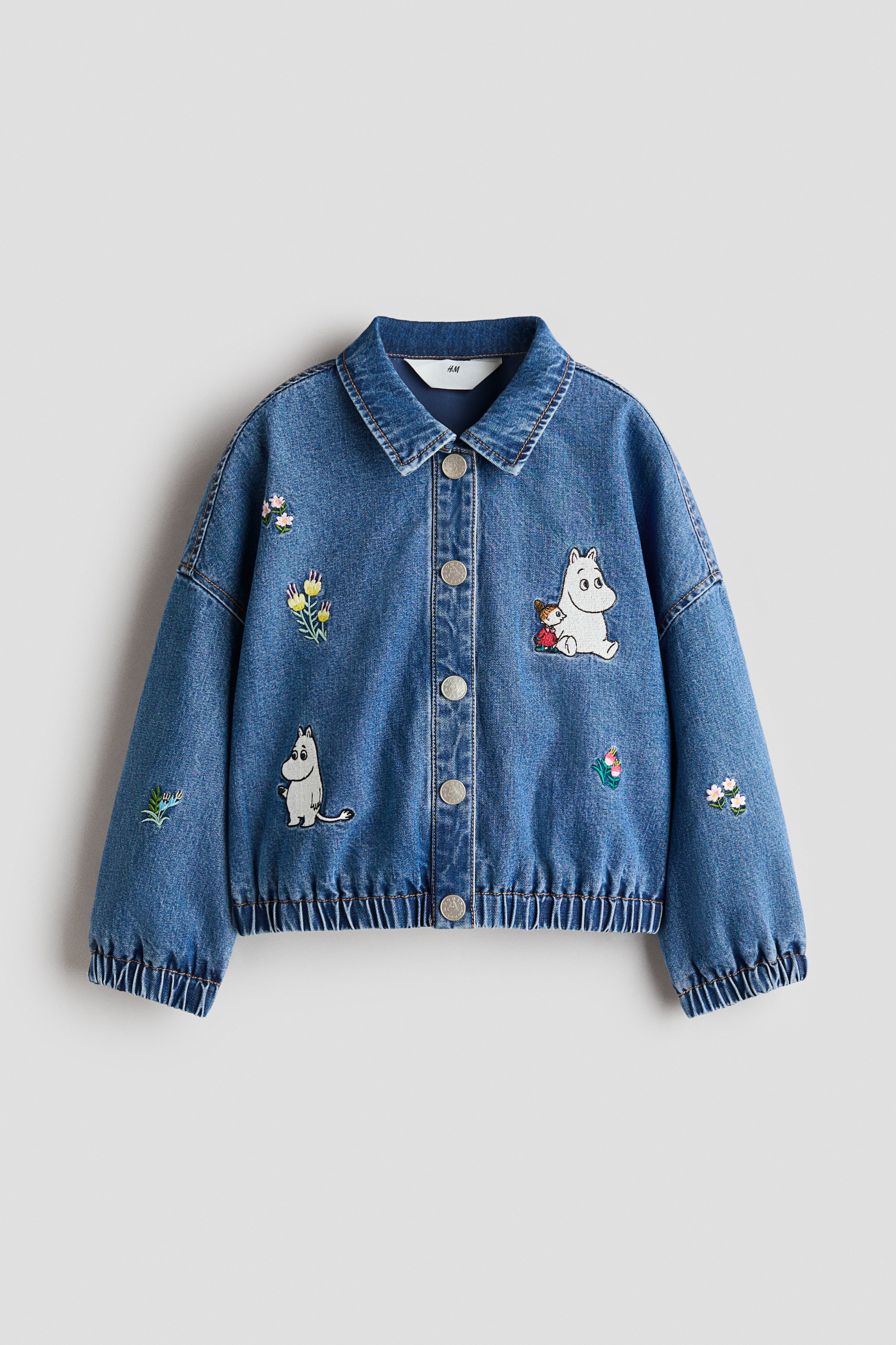 A blue denim jacket is shown from the front, featuring a classic collar, a button-through front and dropped shoulders. The jacket is decorated with embroidered cartoon characters and small flowers on the chest, lower body and sleeves, and is finished with an elasticated hem and cuffs.