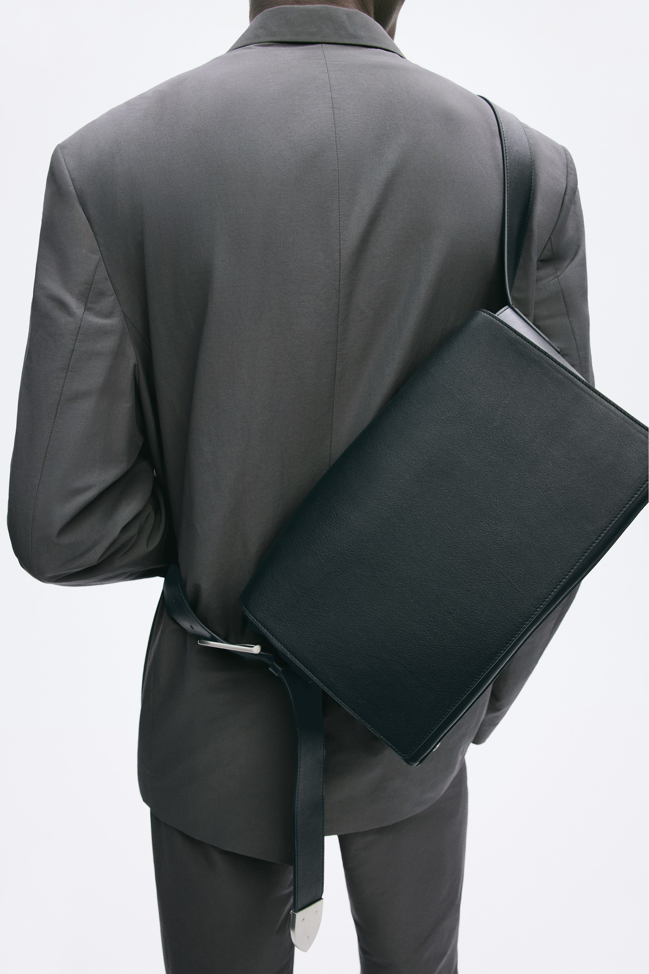 View larger image: A black leather crossbody bag is being worn by the model, featuring a flap and an adjustable shoulder strap with a metal buckle. The bag is positioned across the model’s back, resting against a dark jacket.
