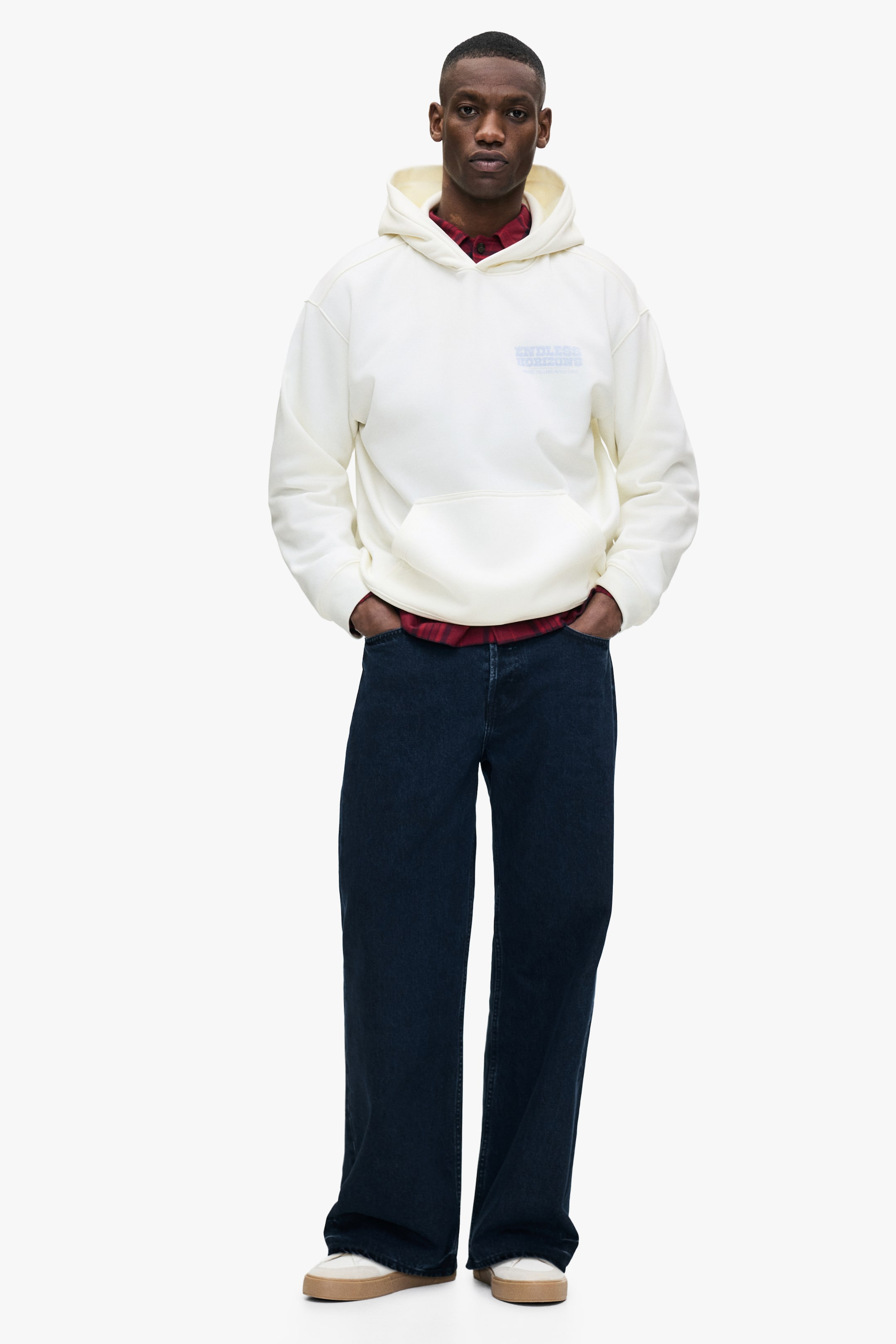 View larger image: A cream hoodie featuring a hood, a front kangaroo pocket, and a light blue text graphic on the chest is being worn, layered over a red plaid shirt with its collar visible.