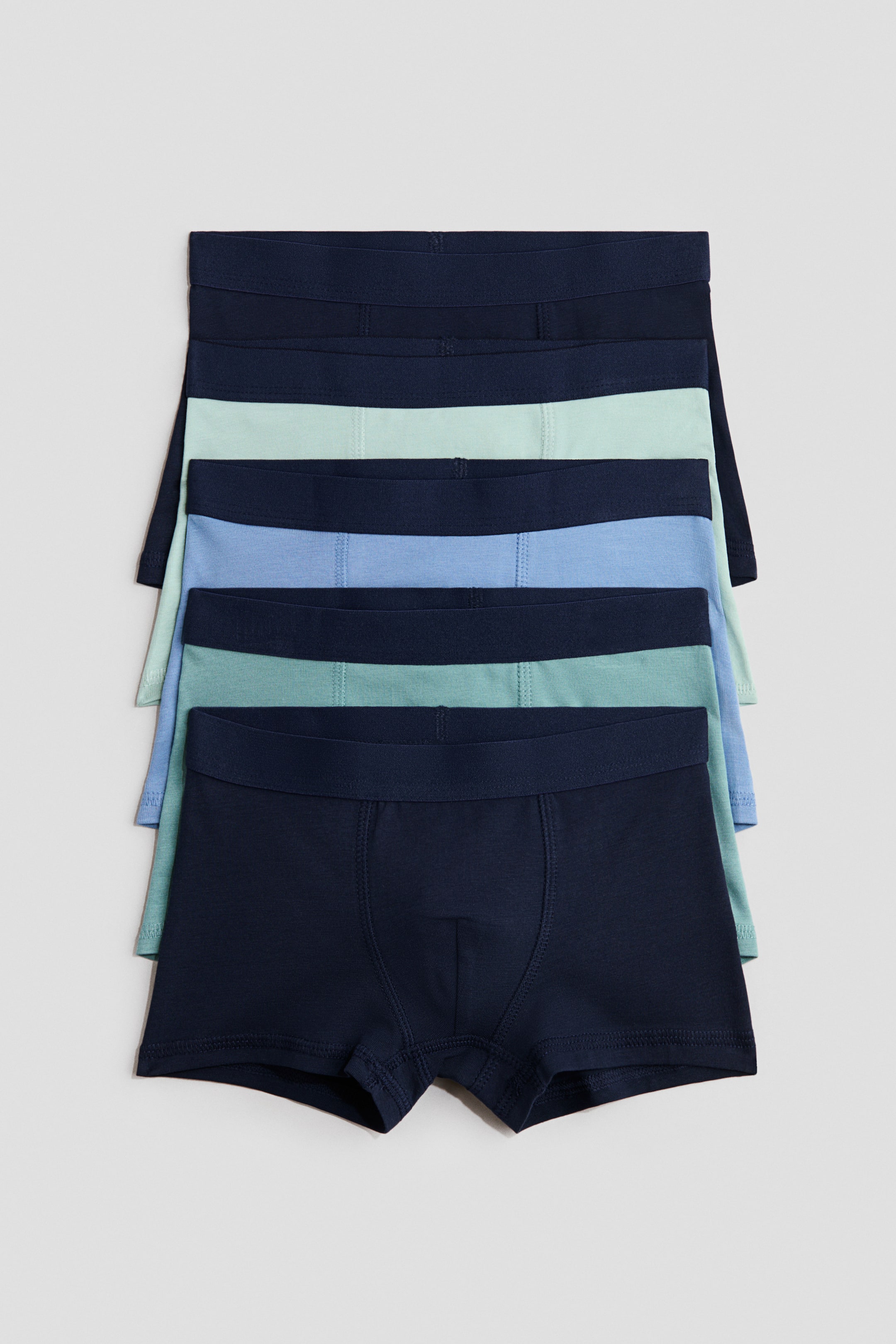 View larger image: Five pairs of trunks crafted from soft cotton jersey, featuring short legs, an elastic waistband, and a lined front, are neatly stacked to showcase their design. The display highlights trunks in a range of dark blue, light blue, and mint green hues.