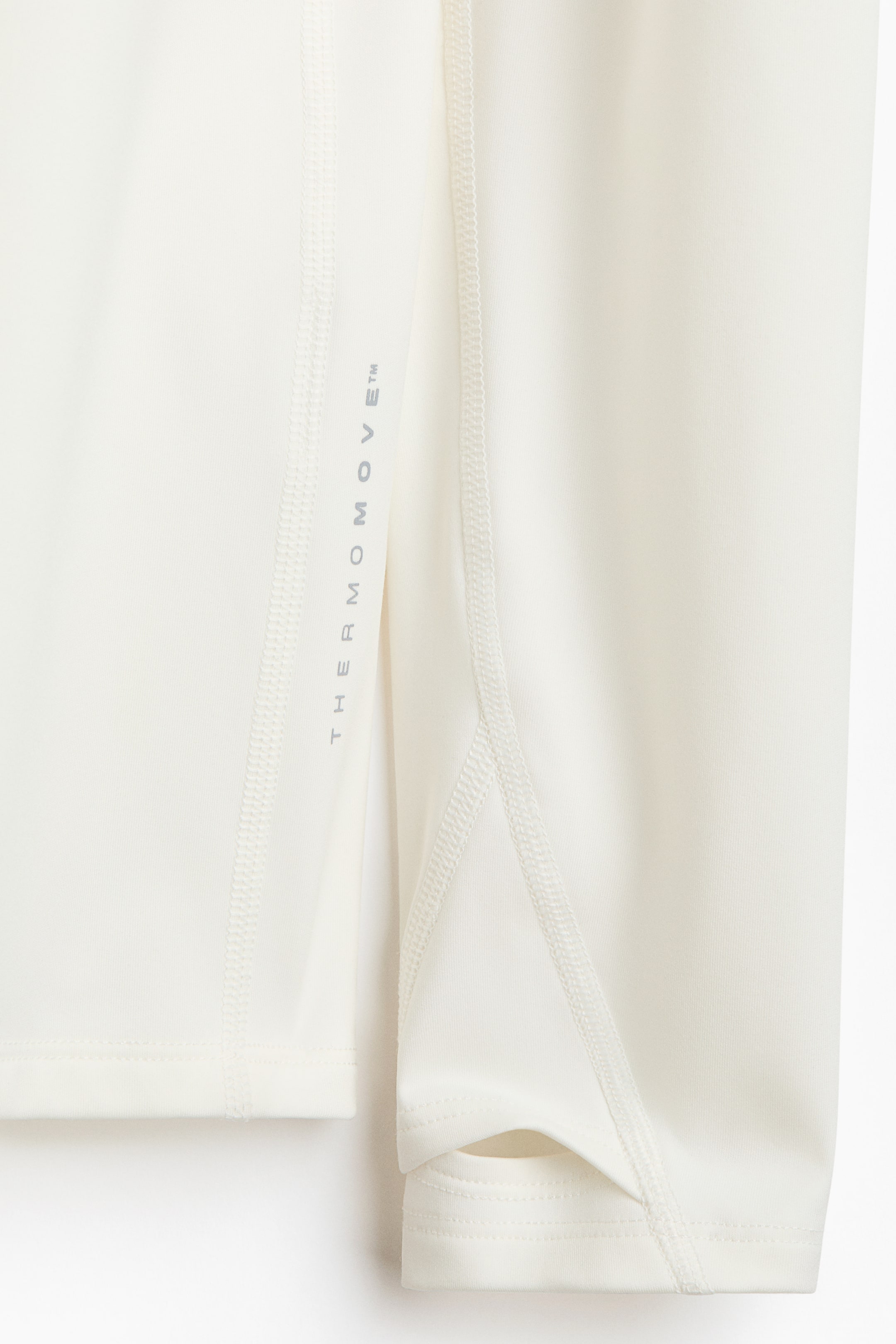 Ampliar la imagen: Una manga de prenda color crema presenta costuras de flatlock, formando paneles a lo largo de su longitud. "THERMOMOVE™" está impreso verticalmente en gris en un panel, y un agujero para el pulgar es visible en el puño.
