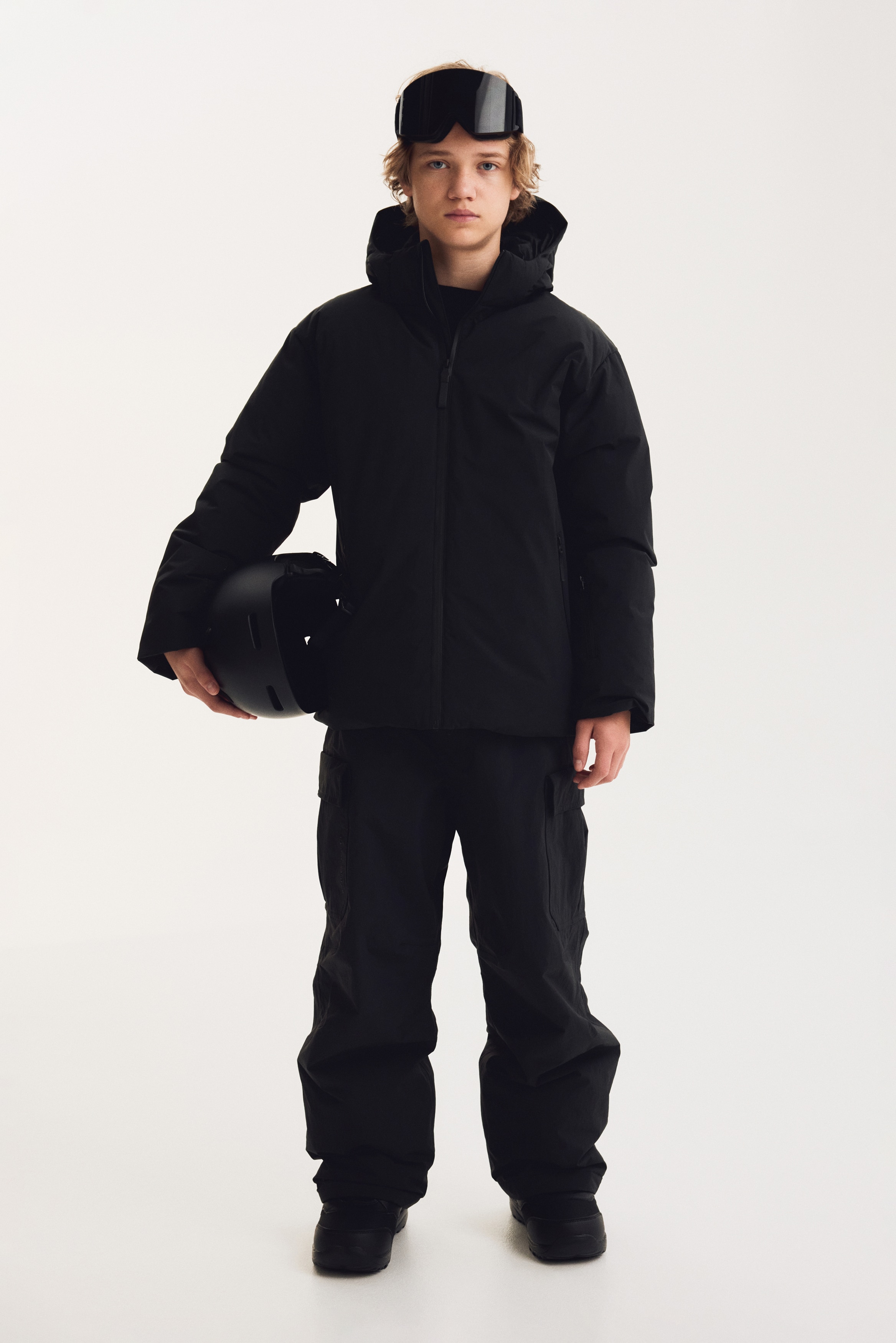 Une veste noire Ã  capuche est prÃ©sentÃ©e de face, avec une coque mate lisse, une fermeture Ã©clair centrale intÃ©grale et une capuche Ã  col montant. La veste prÃ©sente des poches repose-mains zippÃ©es verticales de chaque cÃ´tÃ© et des poignets anti-tempÃªte cÃ´telÃ©s prolongeant les ourlets des manches.