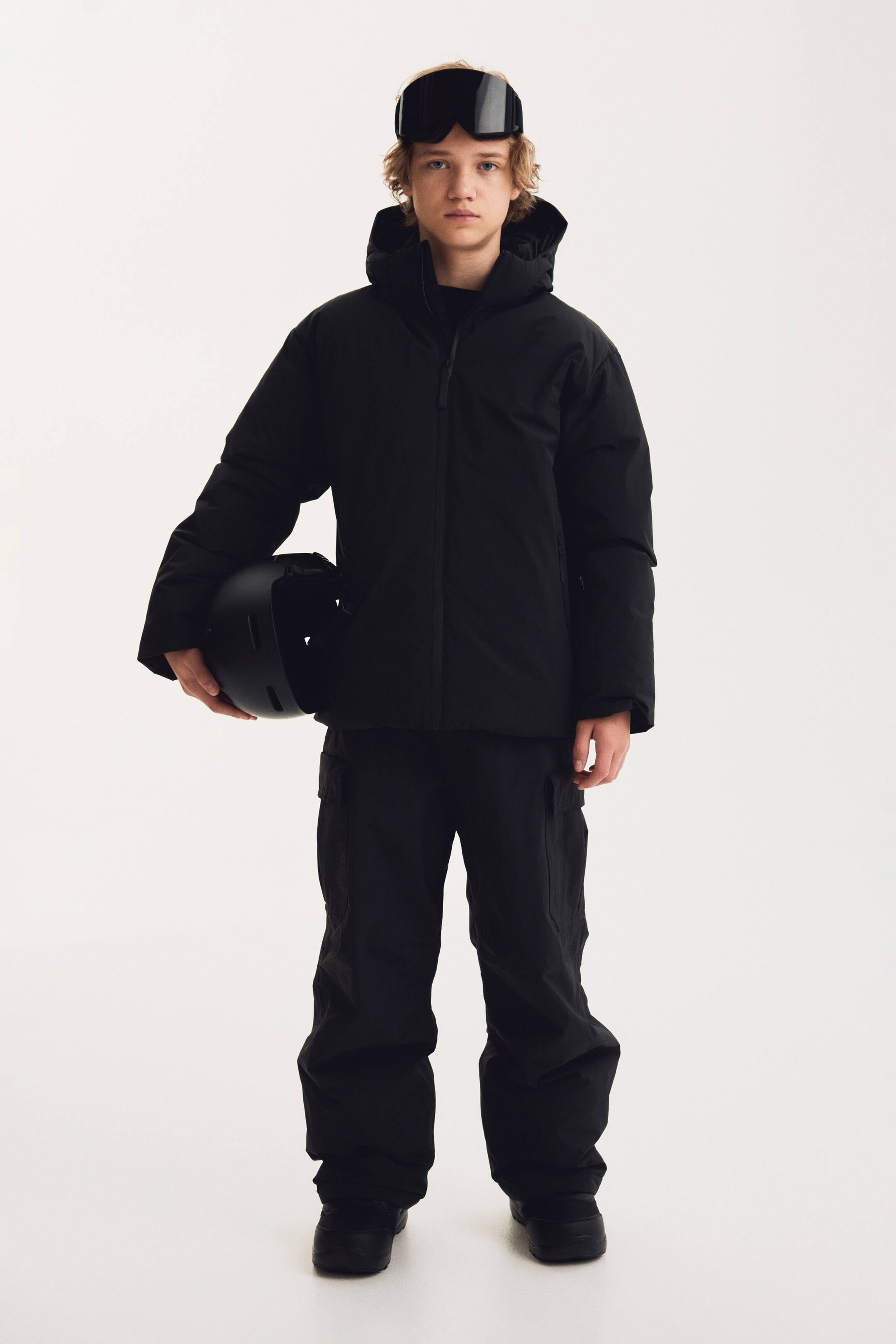 Agrandir l'image: Cette vue de face présente une veste de ski noire à capuche et un pantalon cargo noir assorti. La veste est dotée d’un col montant structuré avec capuche intégrée et d’une fermeture zippée sur le devant, tandis que le pantalon à coupe décontractée affiche des poches cargo latérales.