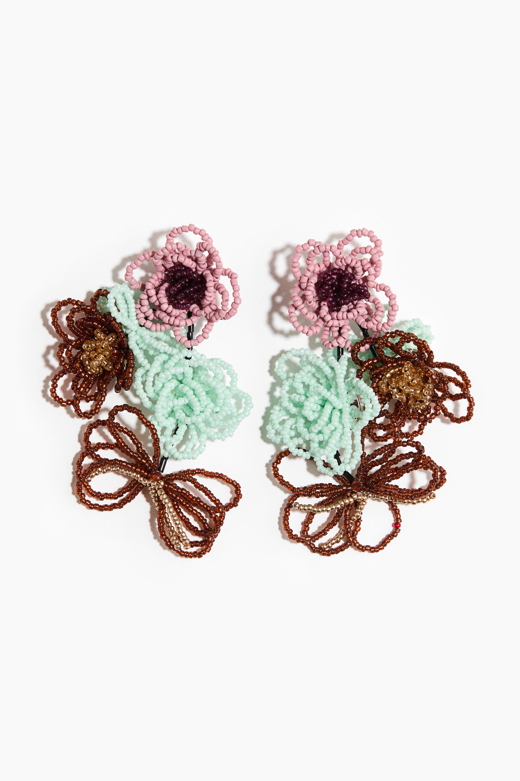 View larger image: The pair of earrings showcases a vertical arrangement of beaded floral and abstract shapes, facing forward. Each earring displays a light pink flower with a dark purple center, a mint green textured cluster, and a brown beaded bow-like shape with gold accents.
