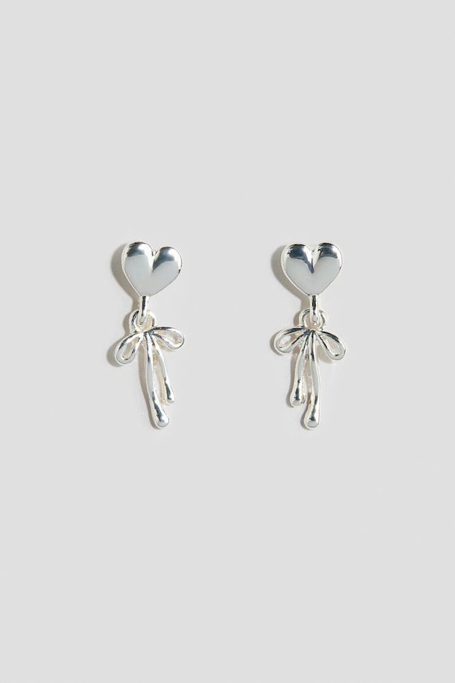 A pair of silver-tone drop earrings is shown from the front, featuring polished, heart-shaped studs. Hanging from each heart is a metallic bow charm with rounded, flowing ribbon ends. - 1