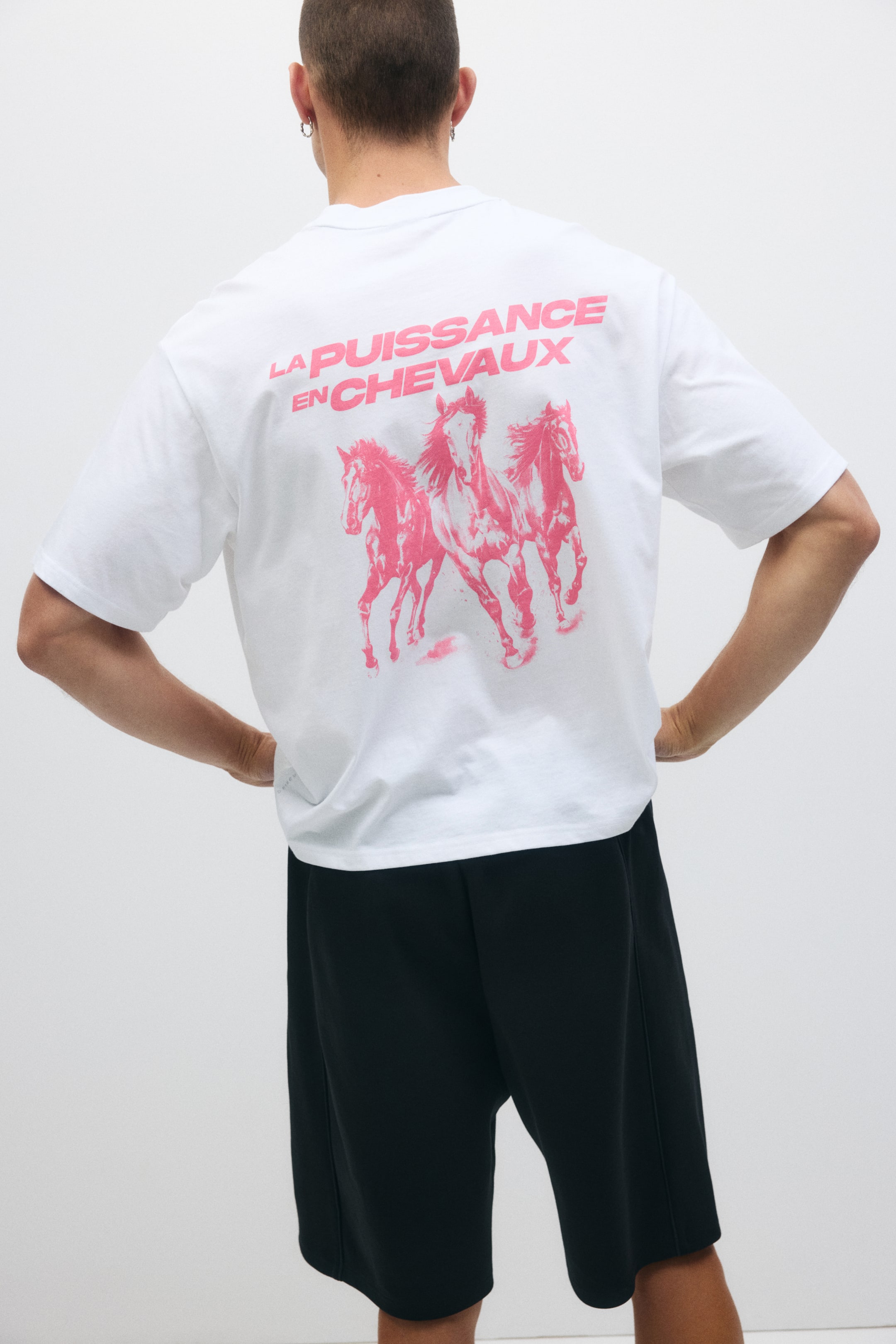 拡大画像を表示: 白いTシャツの背面が映し出されており、大きなピンクのグラフィックには「LA PUISSANCE EN CHEVAUX」というテキストが、3頭の走っている馬の上に描かれています。リラックスフィットの黒いショーツが膝上までの長さで着用されています。
