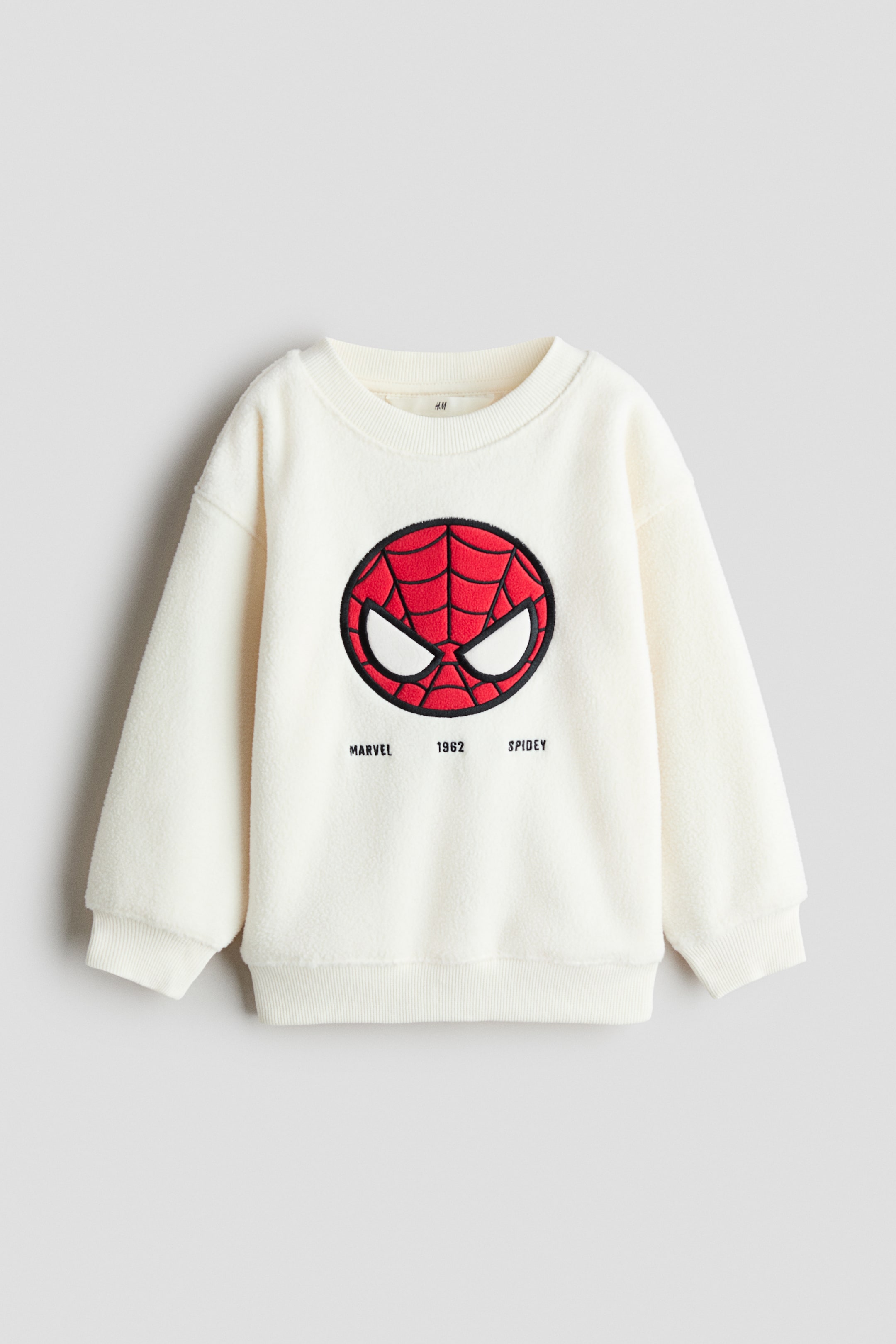 View larger image: An off-white fleece sweatshirt is shown from the front, featuring a ribbed crew neck, dropped shoulders, ribbed cuffs, and a ribbed hem. The chest displays a circular embroidered patch of Spider-Man's face in red, black, and white, with the text "MARVEL 1962 SPIDEY" printed directly below.