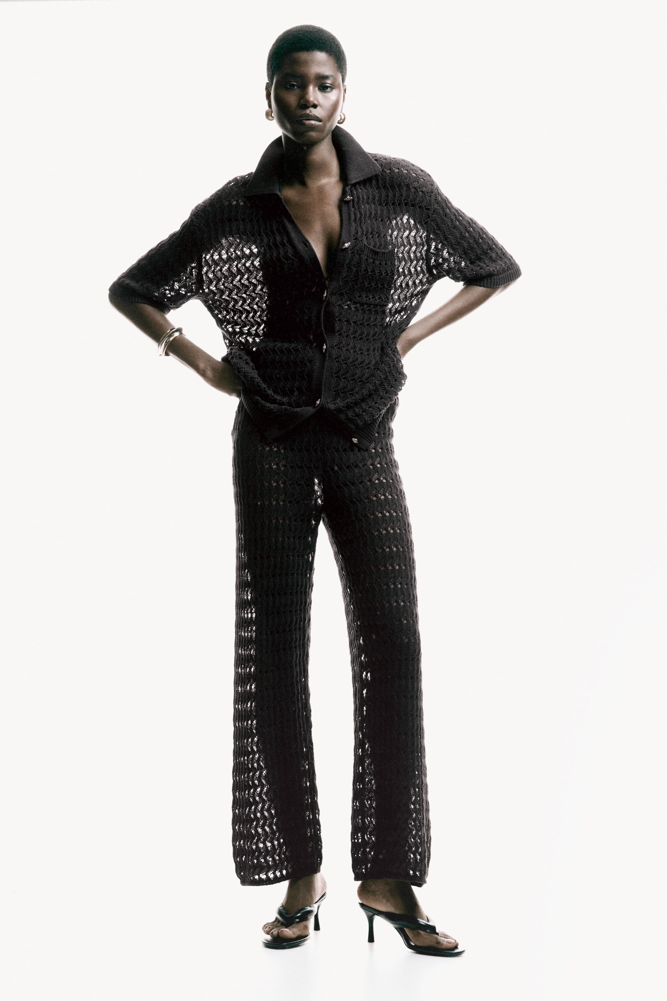 View larger image: A black open-knit crochet short-sleeved shirt, featuring a collar, button-front closure, and a single chest pocket, is showcased with matching wide-leg pants and black high-heeled thong sandals, all facing forward.