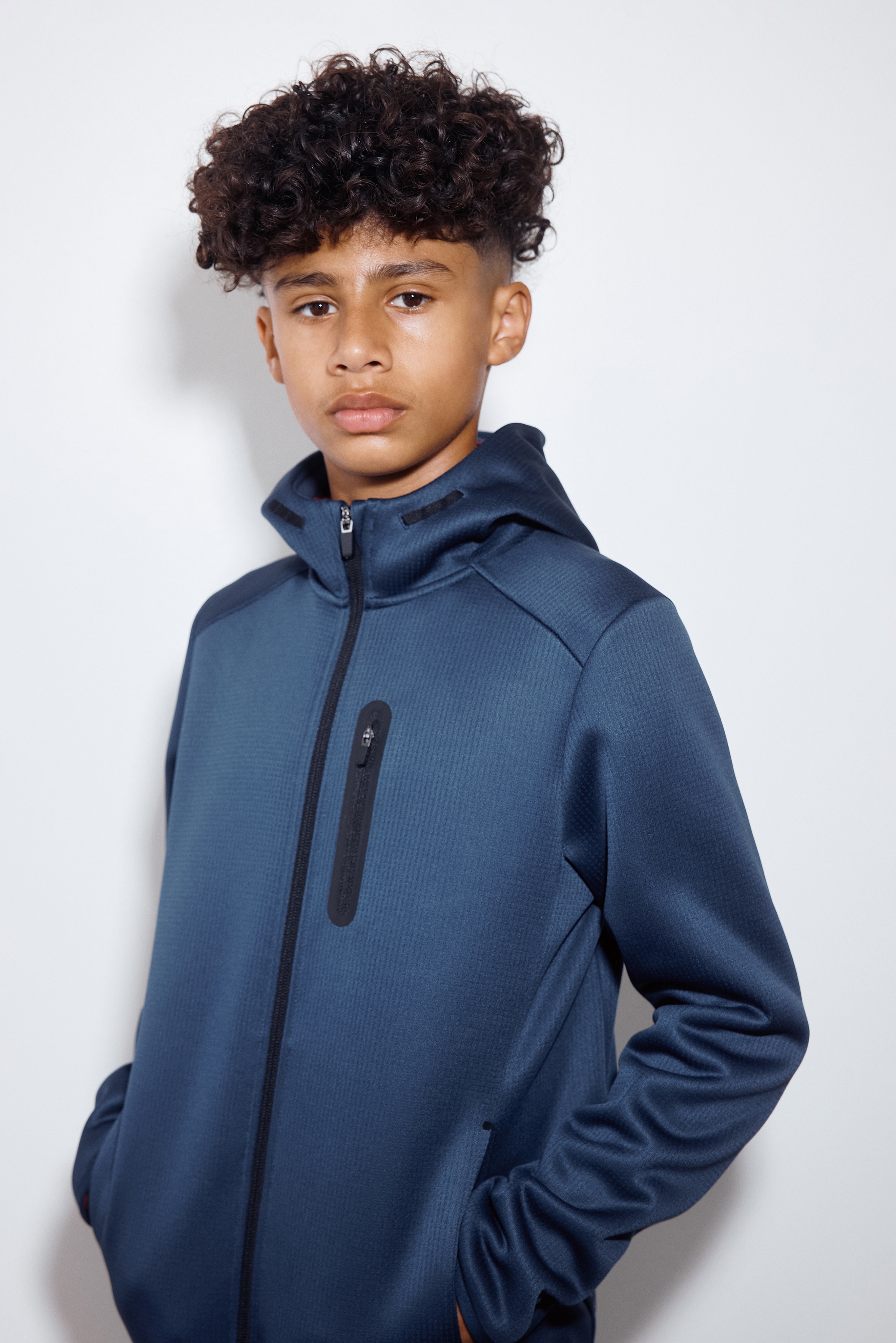 A blue zip-up hooded jacket is shown from the front, featuring a textured, finely ribbed fabric and a full-length black front zip. It is designed with an attached hood, a vertical zip-pocket on the left chest with black bonding, and long sleeves with neat, narrow cuffs.