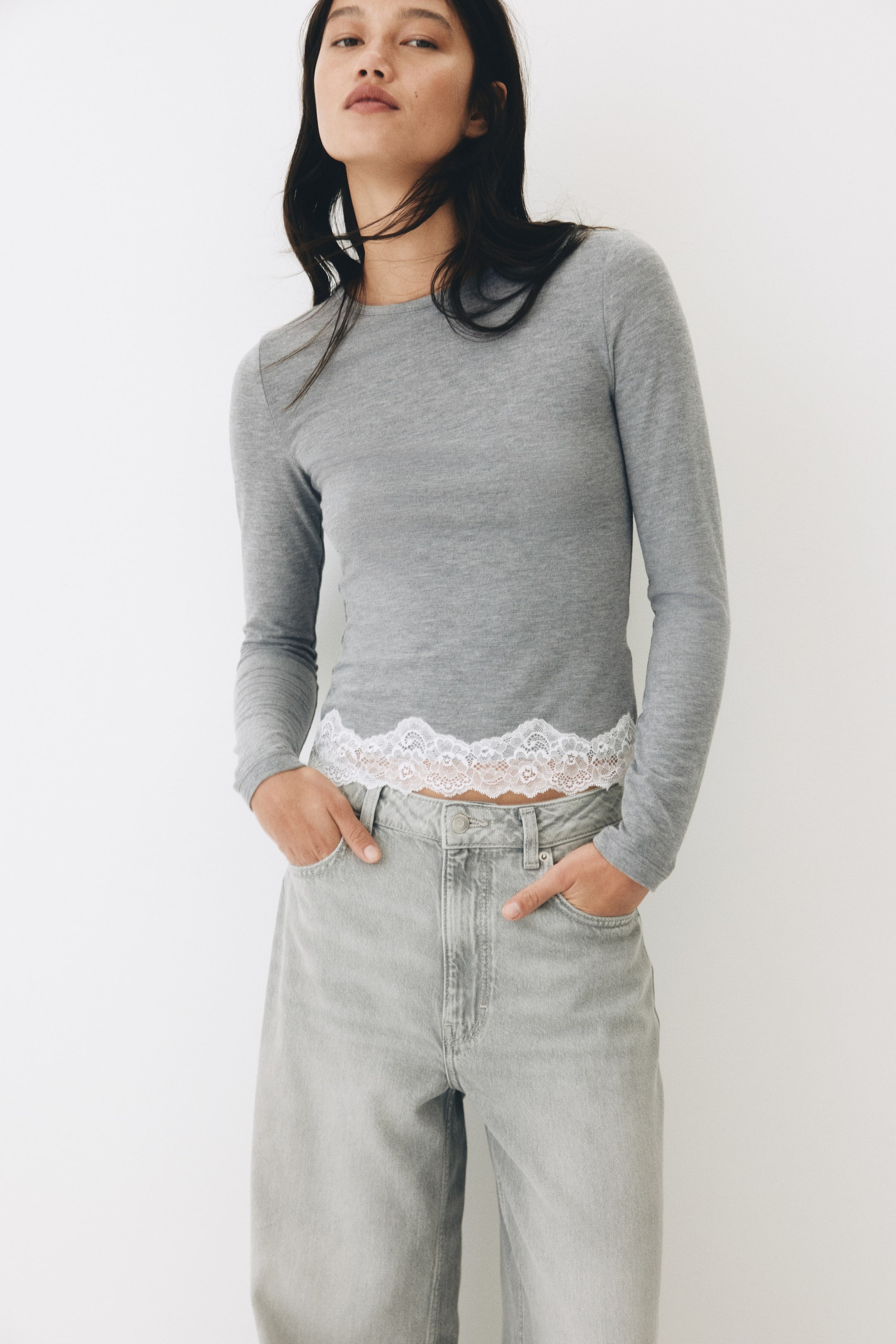 View larger image: The model is wearing a fitted, long-sleeve top in soft gray jersey featuring a round neckline and a white lace-trimmed hem. Standing with hands in the pockets of light gray jeans, the model prominently showcases the top's lace hemline.