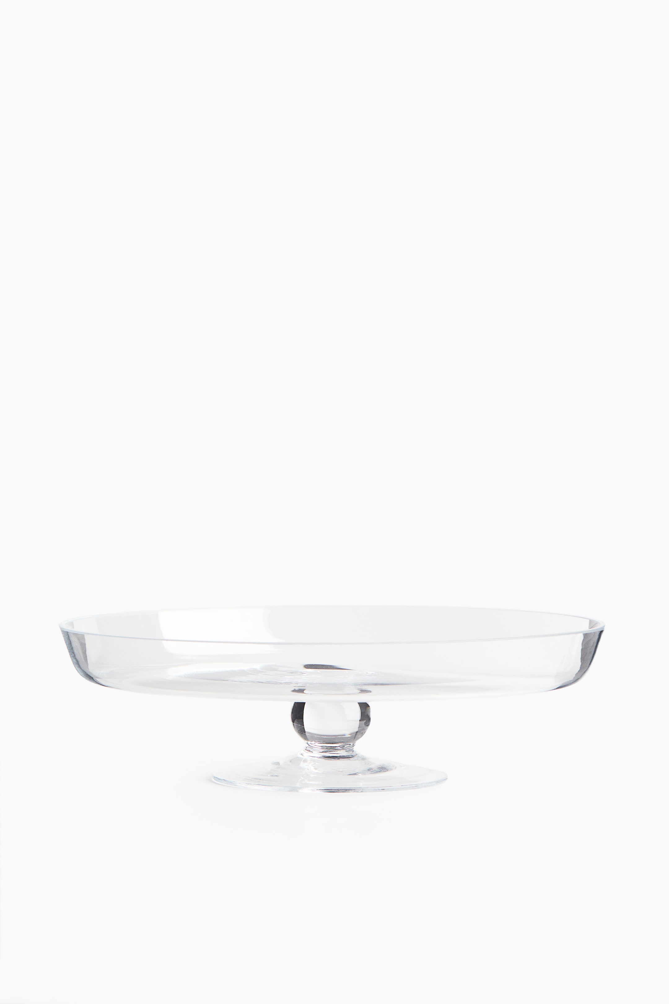 View larger image: A clear glass pedestal stand is resting on a flat surface, facing forward. It features a wide, shallow circular dish with a slightly raised rim, supported by a short stem with a spherical detail and a flared circular base.